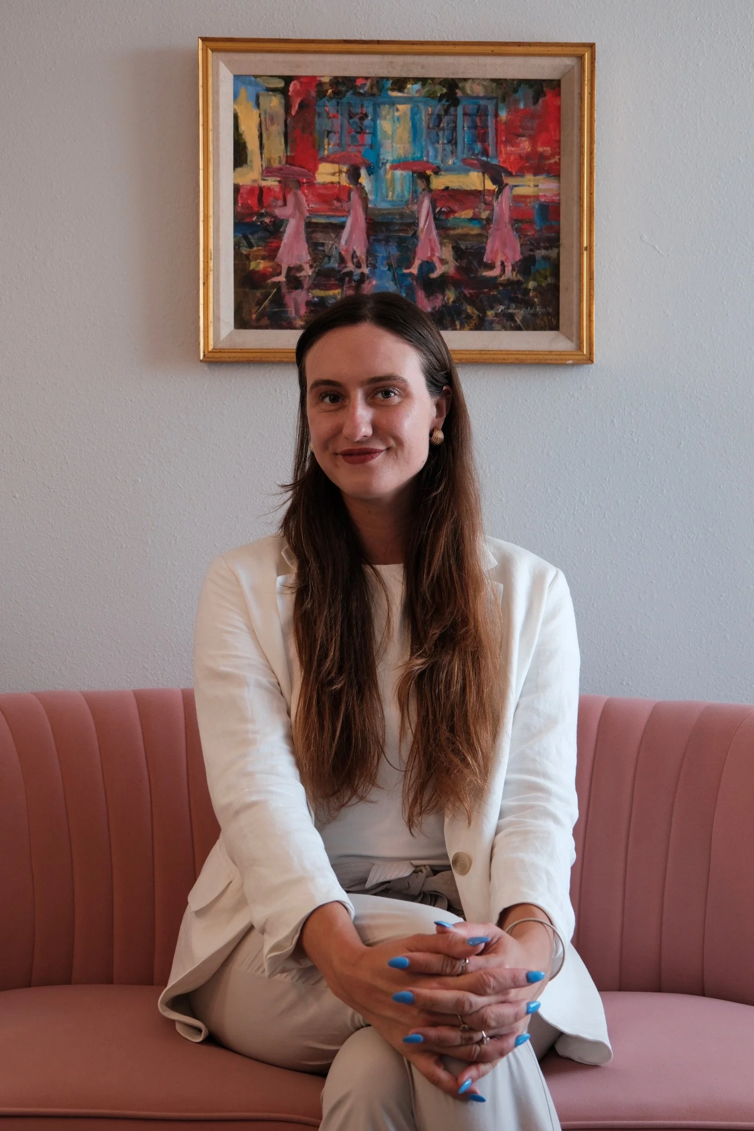 A woman sitting on a pink velvet sofa with her hands clasped, wearing a white blazer and beige pants, with long brown hair and blue nails, in front of a white wall with a colorful painting of women holding umbrellas.