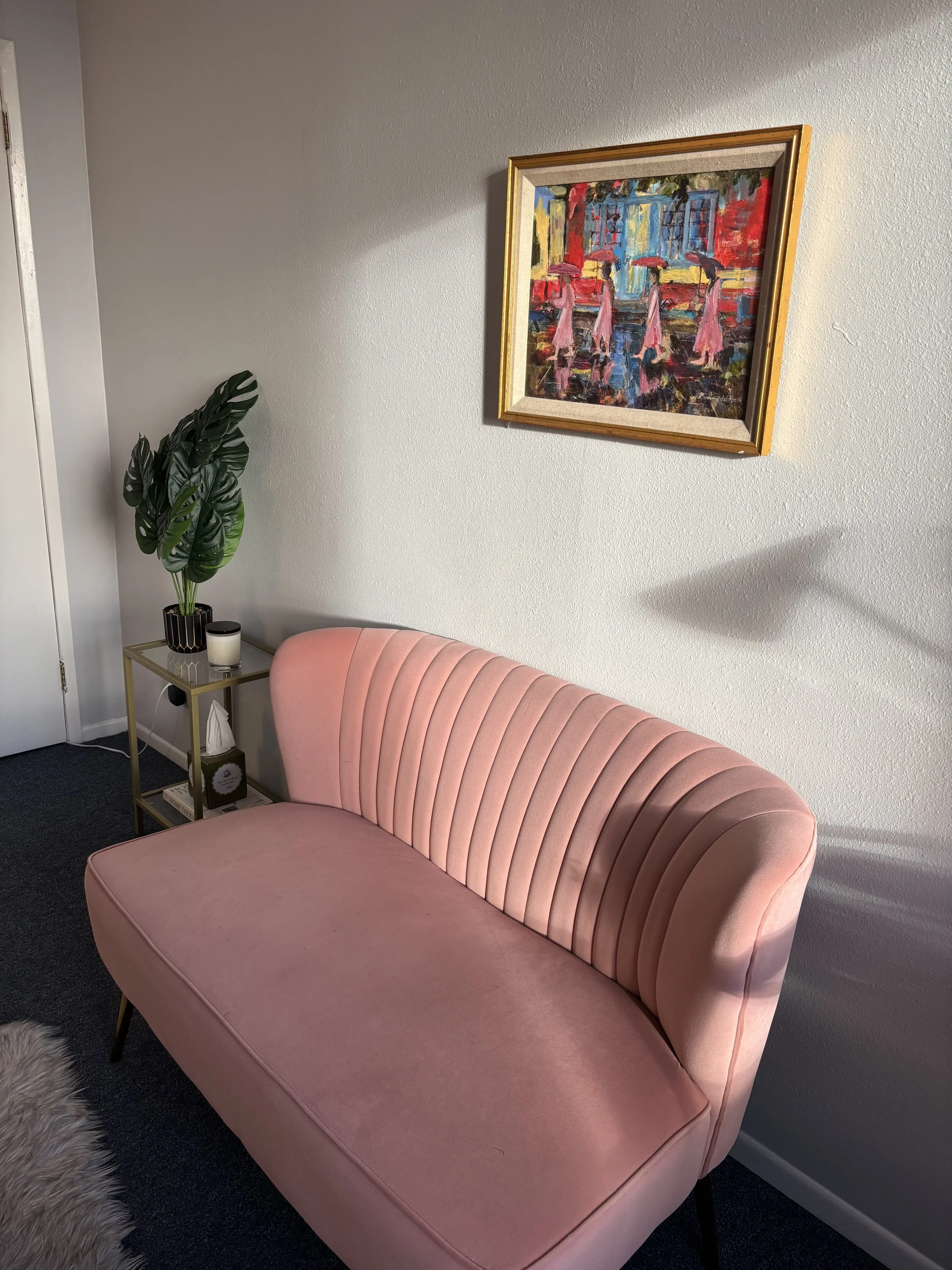 A pink velvet sofa with vertical tufting is placed against a light gray wall. To the left, a small decorated side table holds a potted plant with large green leaves, a candle, and some books. A colorful abstract painting with red, blue, yellow, and pink tones hangs on the wall above the sofa.