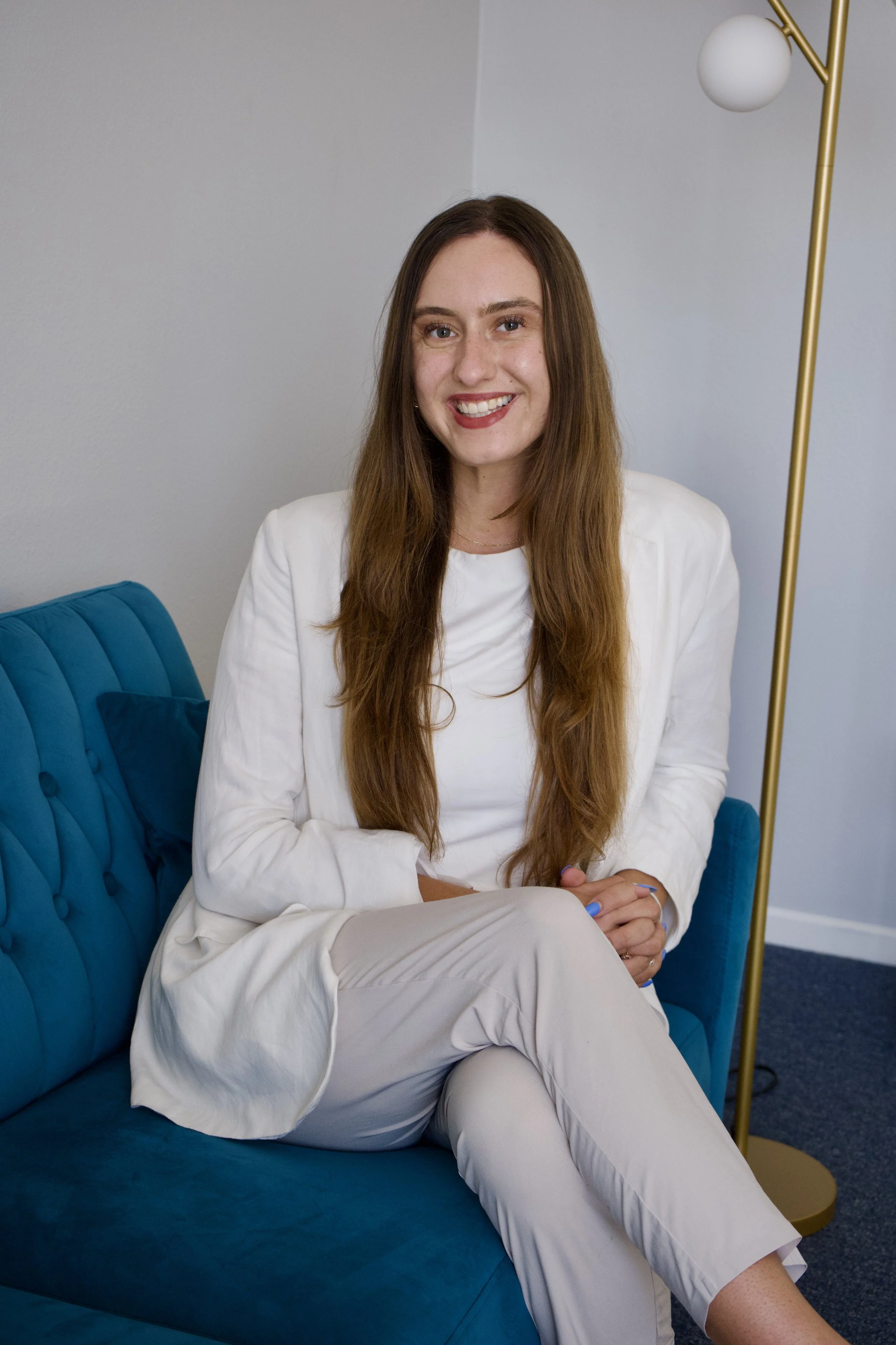 A woman with long brown hair wearing a white outfit, sitting on a blue sofa, smiling, with a brass floor lamp and gray walls in the background.
