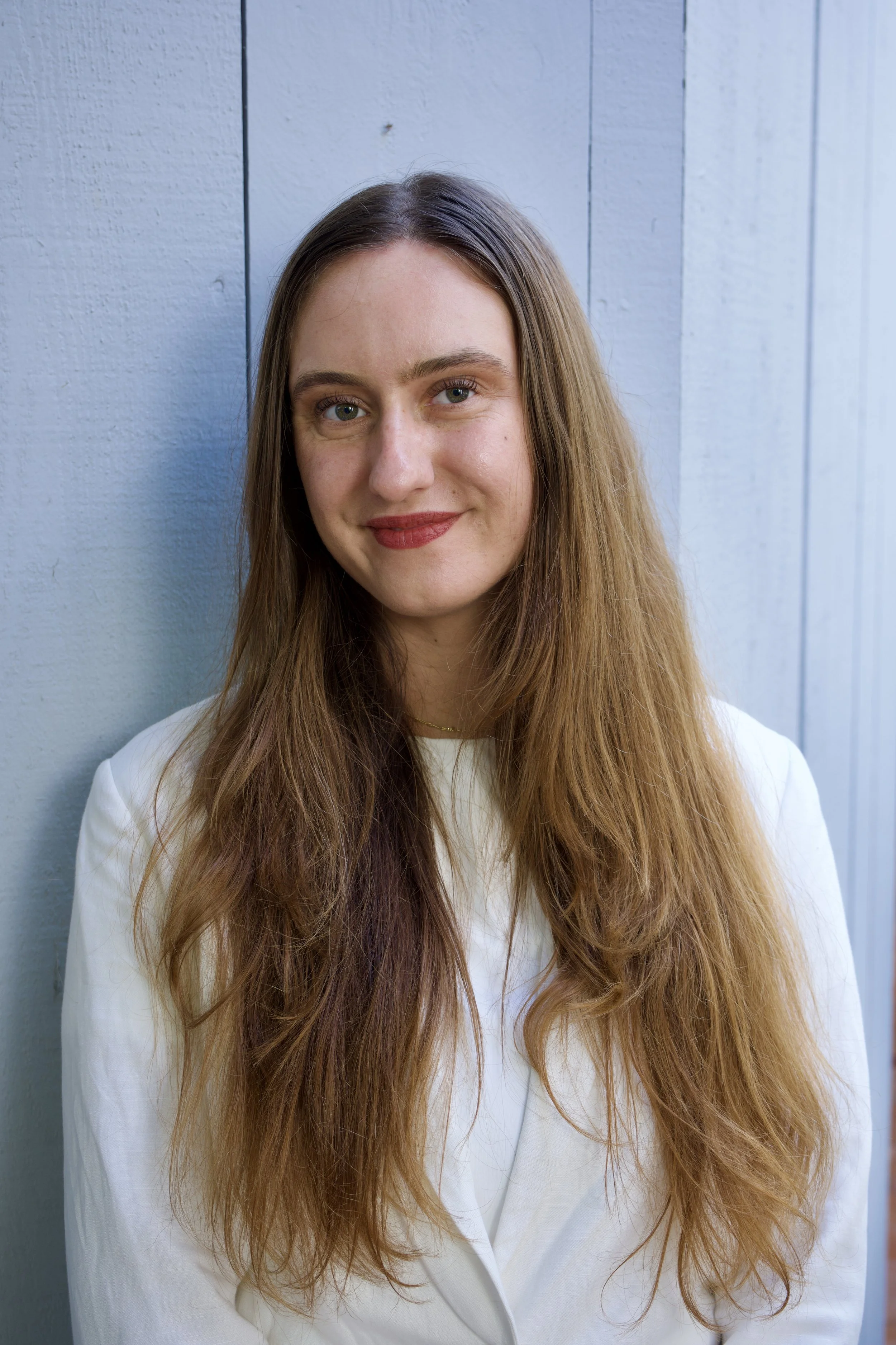 A woman with long, wavy red hair, blue eyes, and light skin, smiling slightly, standing in front of a light blue wooden wall.
