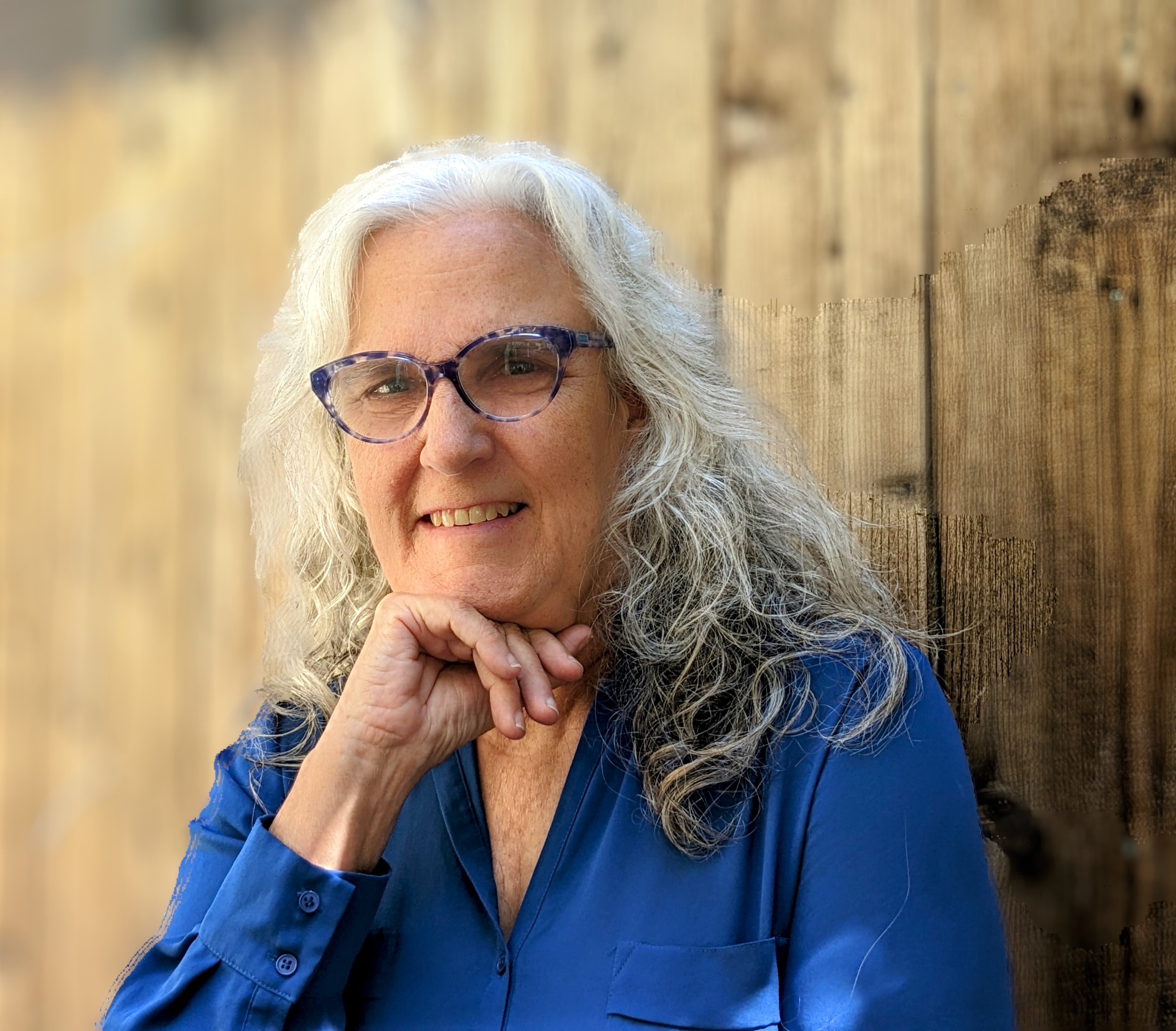 A woman with gray hair and glasses smiling, resting her chin on her hand, standing in front of a wooden fence.