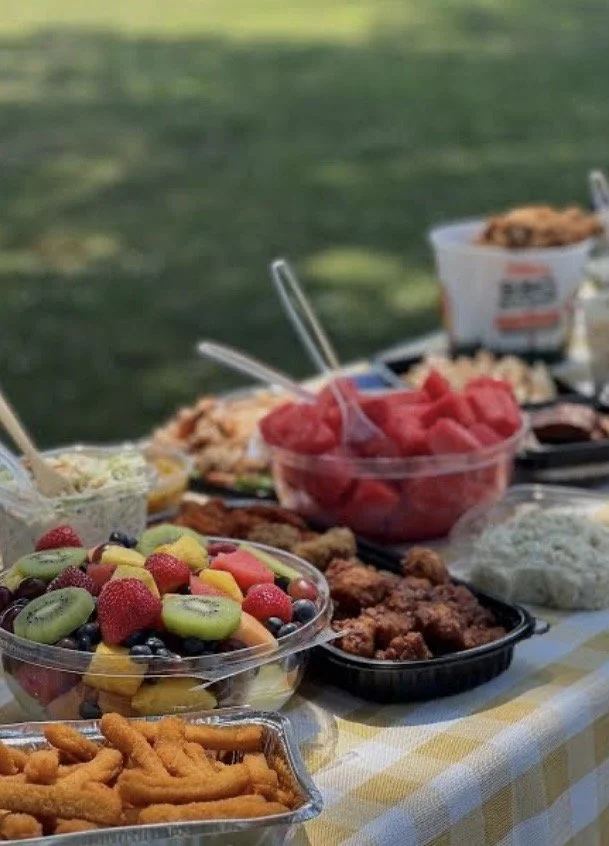 Assorted picnic food on a yellow checkered tablecloth including fruit salad, watermelon, fried chicken, pasta salad, and other dishes in containers, outdoors with green grass in the background.