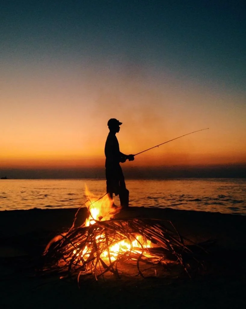 Silhouette of a person fishing at sunset on a beach, with a campfire in the foreground and the ocean in the background.
