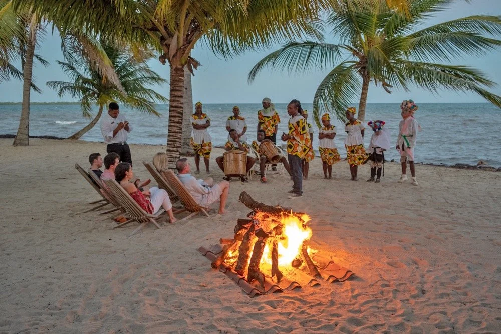 People sitting in chairs and standing around a bonfire on the beach under palm trees, with musicians and dancers in traditional African attire performing in the background.