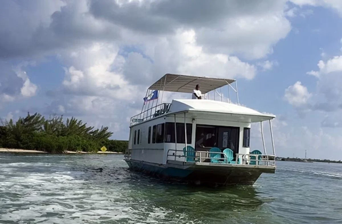 A white boat with a shaded upper deck and turquoise chairs on the lower deck is moving across the water under a partly cloudy sky.