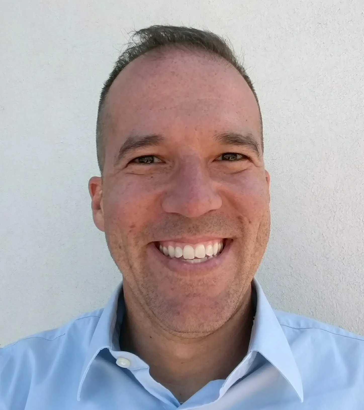 Close-up of a smiling man with short dark hair, wearing a light blue collared shirt, standing against a plain, light-colored wall.