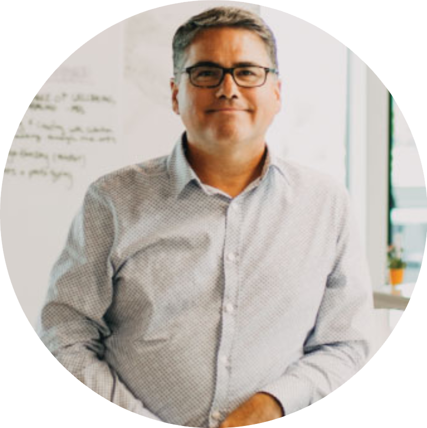 A man named Ray DePaul with glasses and short hair standing in an office, smiling, with a whiteboard and window in the background.