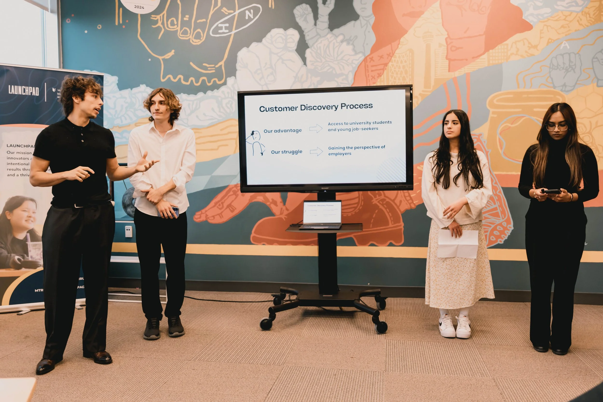 A group of  young adults presenting in front of a colorful mural. A large screen displays a presentation slide titled 'Customer Discovery Process,' with bullet points and simple illustrations. The presenter on the left is speaking and gesturing with