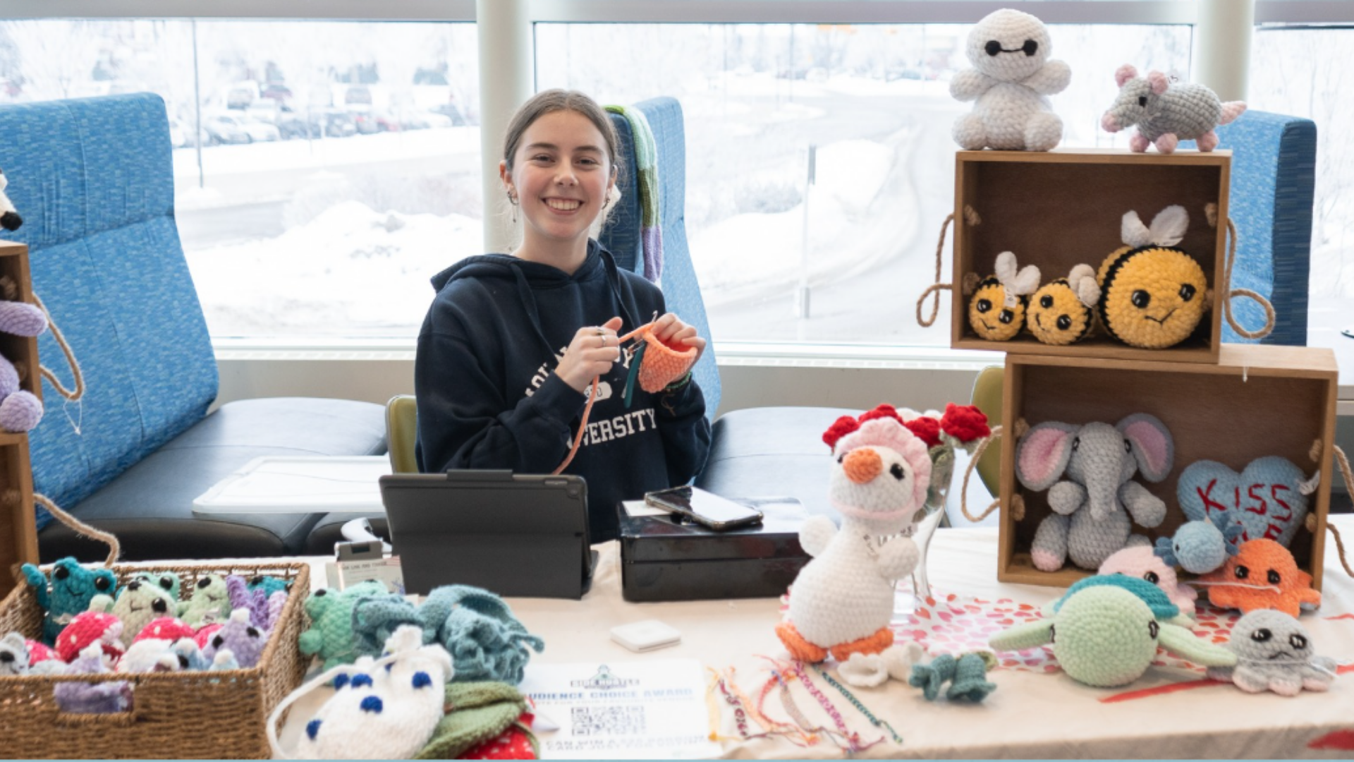 Young woman with brown hair smiling while knitting crochet toys at a craft fair booth, surrounded by handmade stuffed animals and toys such as bees, elephants, and ducks.