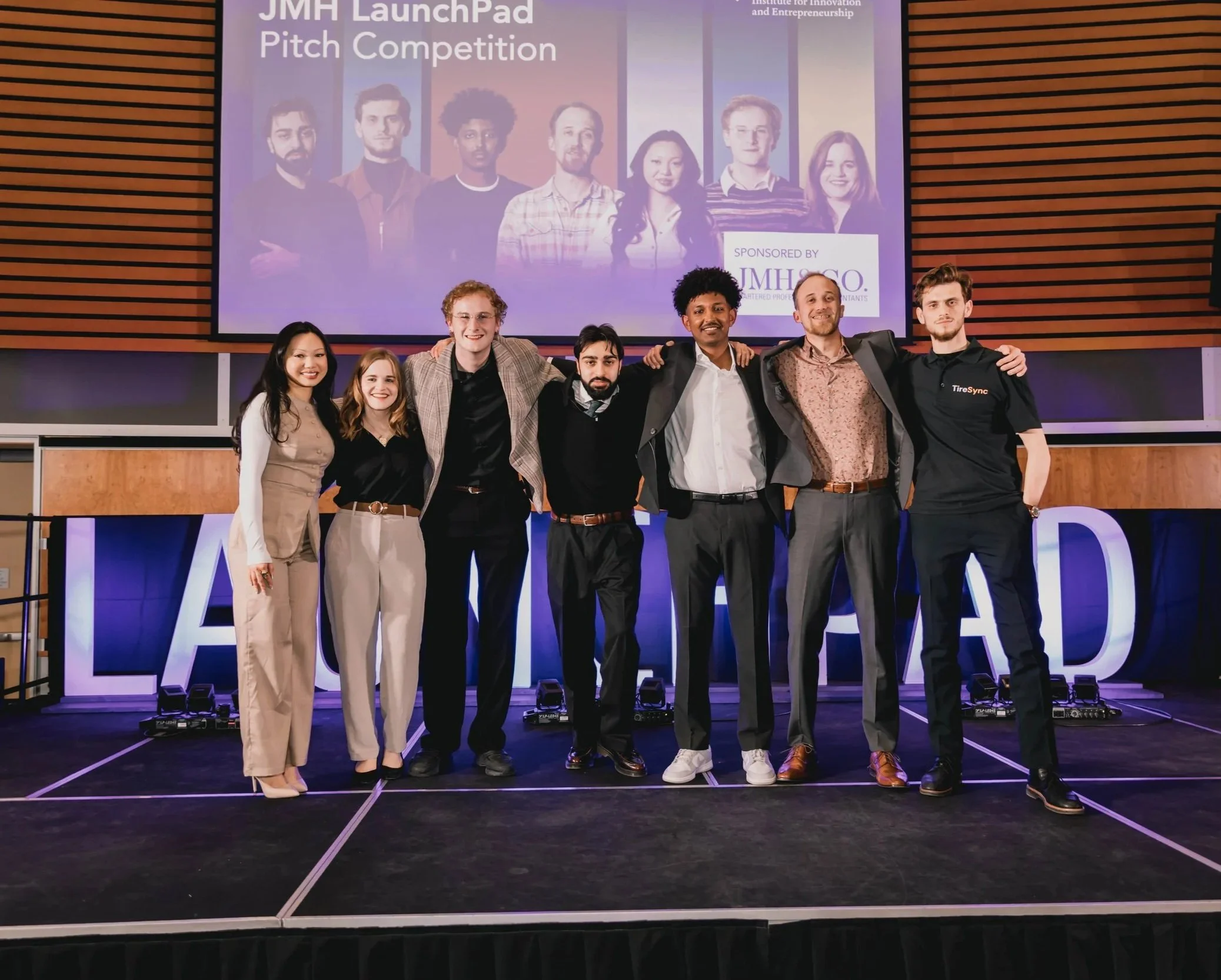 Group of seven diverse young students standing together on stage at the JMH LaunchPad Pitch Competition, with large screen behind them.