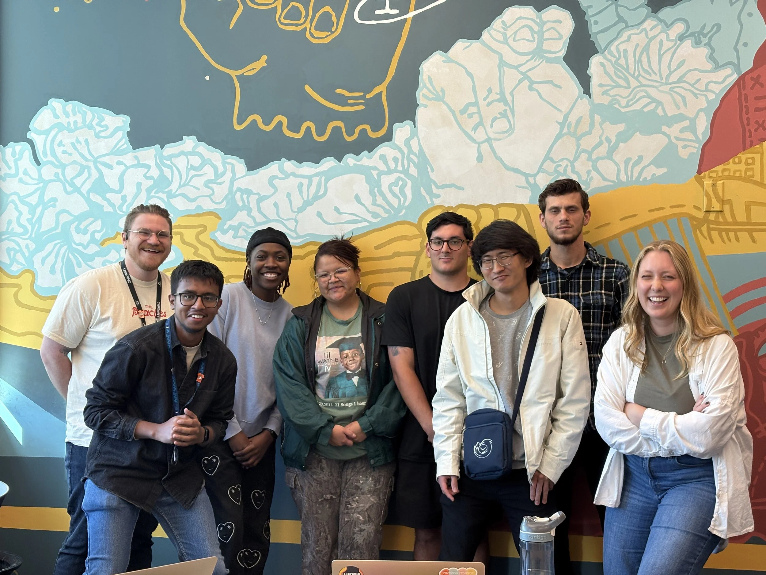 A group of nine young adults standing together in front of a colorful mural in the Slate Innovation Lab, smiling and posing for the photo.