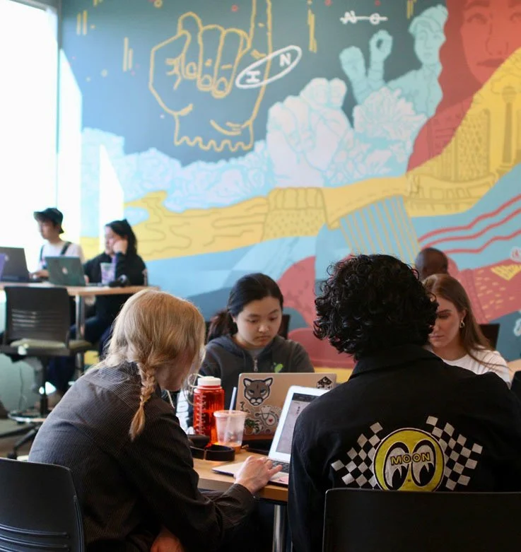 A group of young women sitting at a table in Slate Lab using laptops. A large, vibrant mural with abstract art decorates the wall behind them.