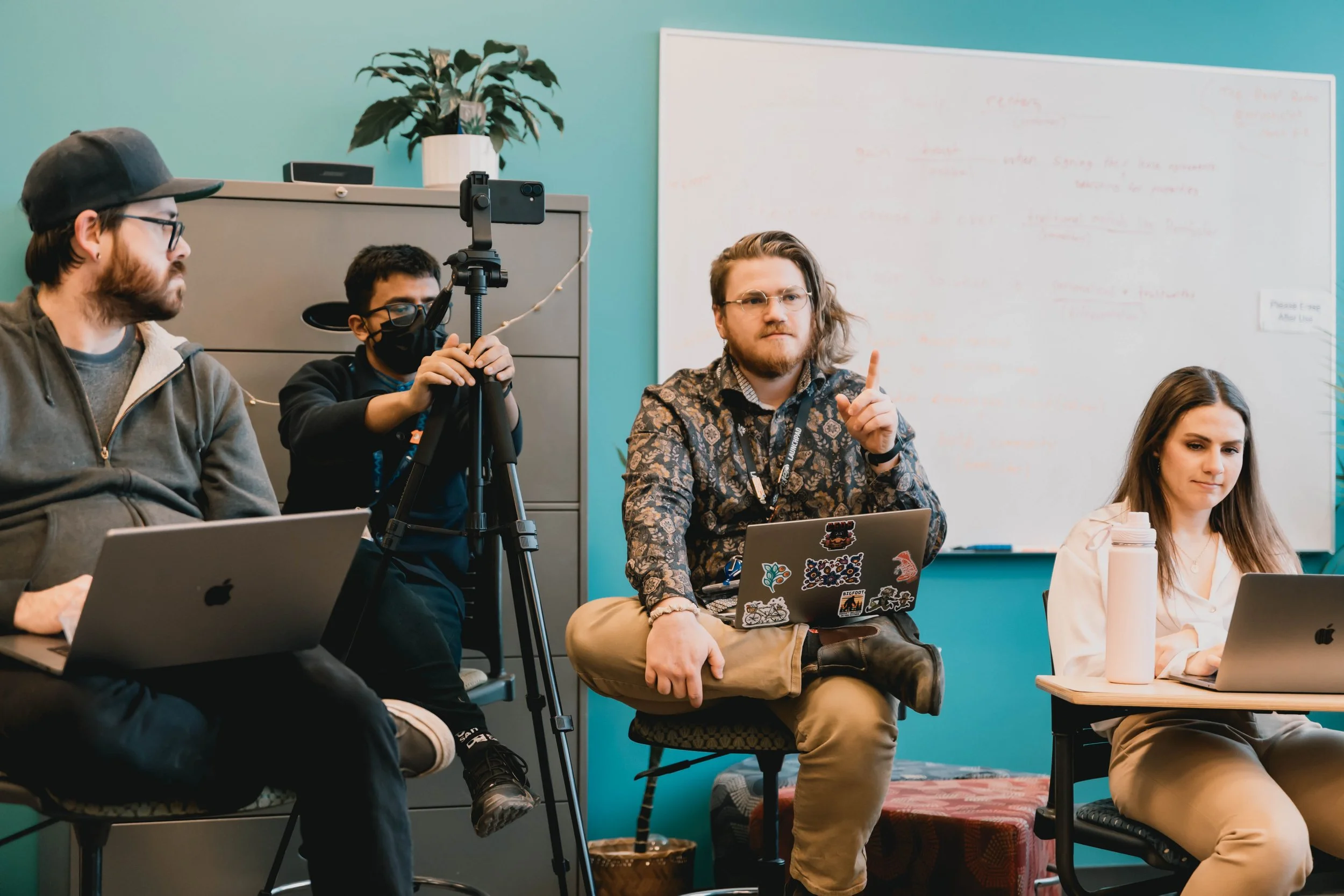 Group of students and staff in a classroom or meeting room, some wearing glasses, one with a mask, sitting with laptops, one person taking a photo or recording with a smartphone on a tripod, a whiteboard in the background.