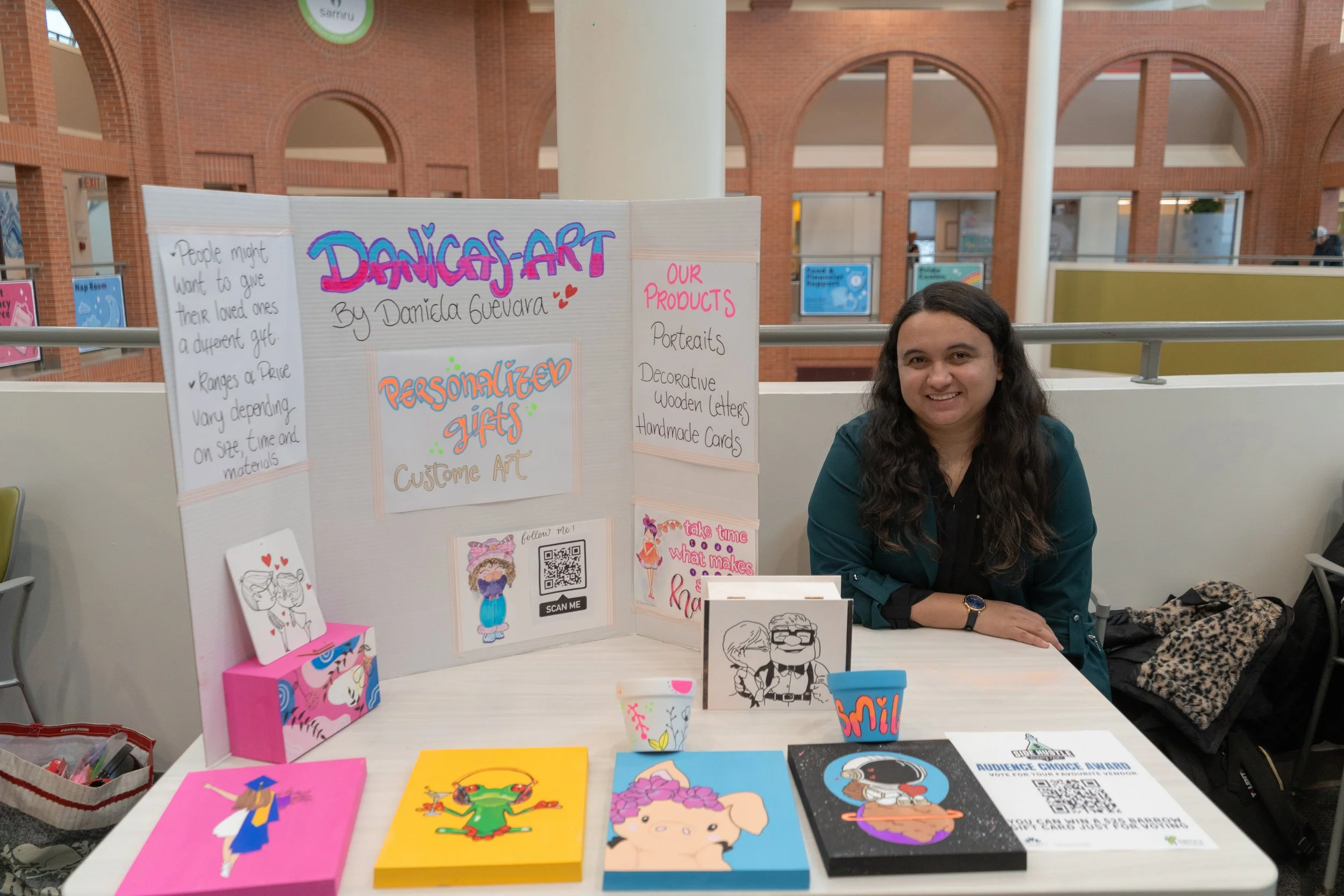 Young woman with brown hair smiling while knitting crochet toys at a craft fair booth, surrounded by handmade stuffed animals and toys such as bees, elephants, and ducks.