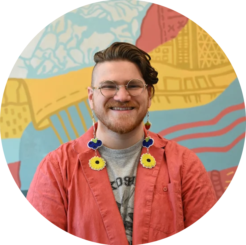 A man named Orin Bailey Davies with long brown hair, glasses, and a beard smiling, wearing a red shirt, colorful flower earrings standing in front of a colorful, abstract mural.