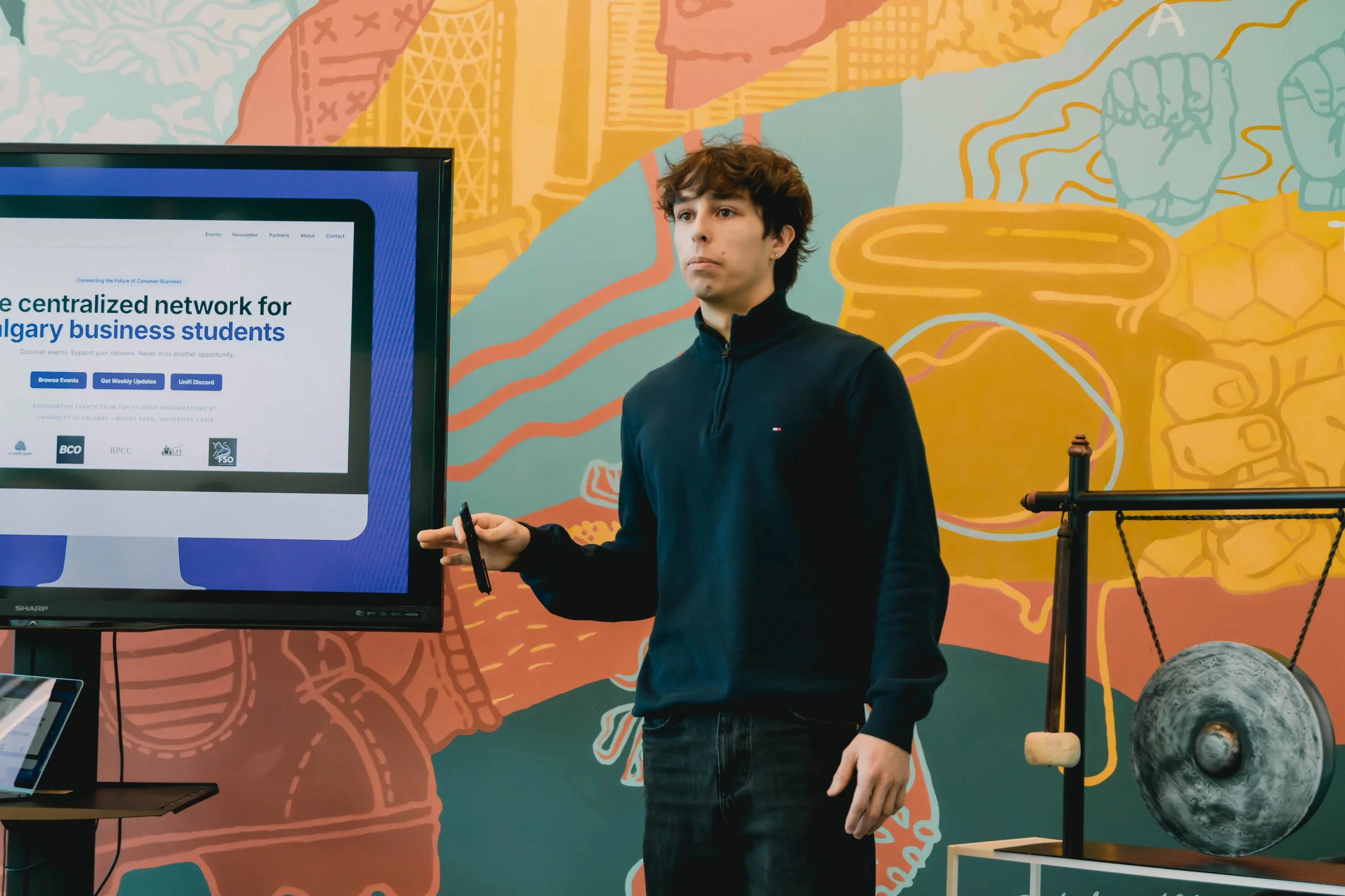 A young man standing next to a large monitor displaying a website about connecting Canadian business students. The background features a colorful mural. The young man holds a pointer and is presenting.