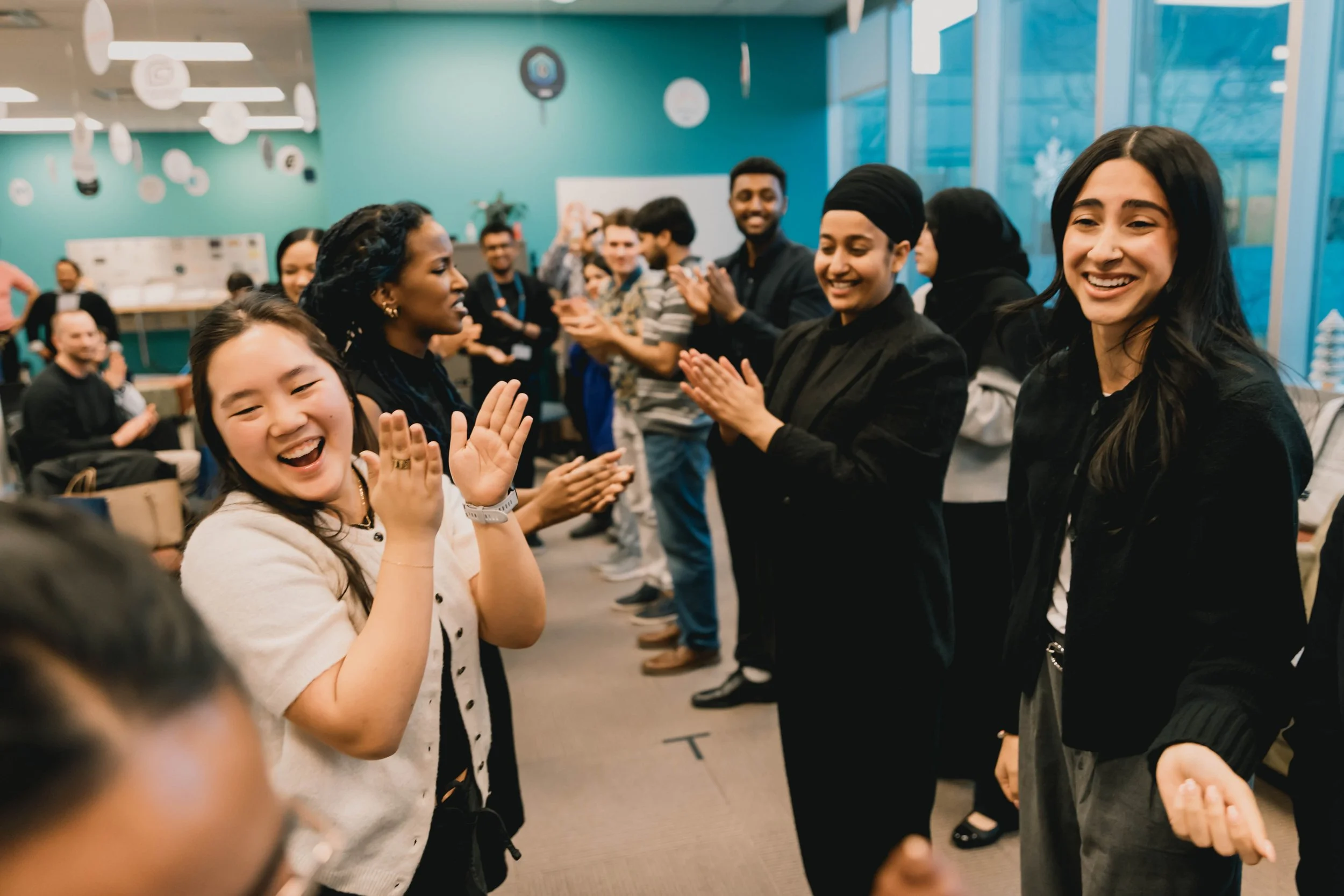 Group of people, including women and men of various ethnicities, standing in a line and clapping, smiling, and enjoying an indoor event.