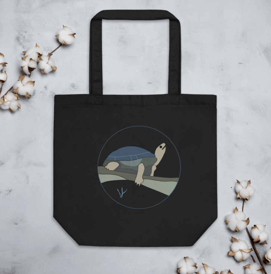 Black tote bag with a turtle illustration on it, lying on a light gray surface surrounded by cotton branches.