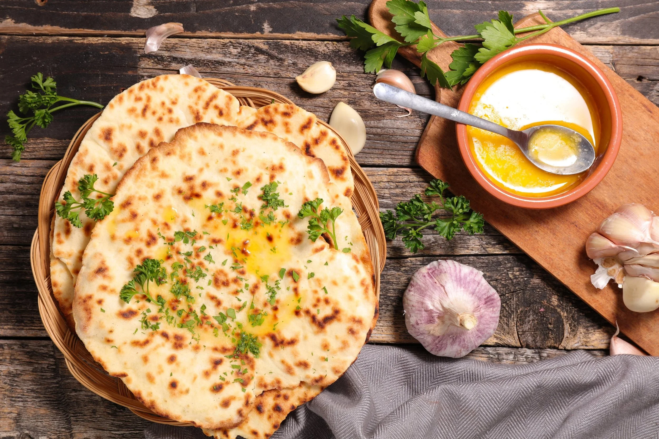 A basket of naan bread garnished with chopped herbs surrounded by garlic, parsley, and a bowl of eggs and spices on a rustic wooden surface.