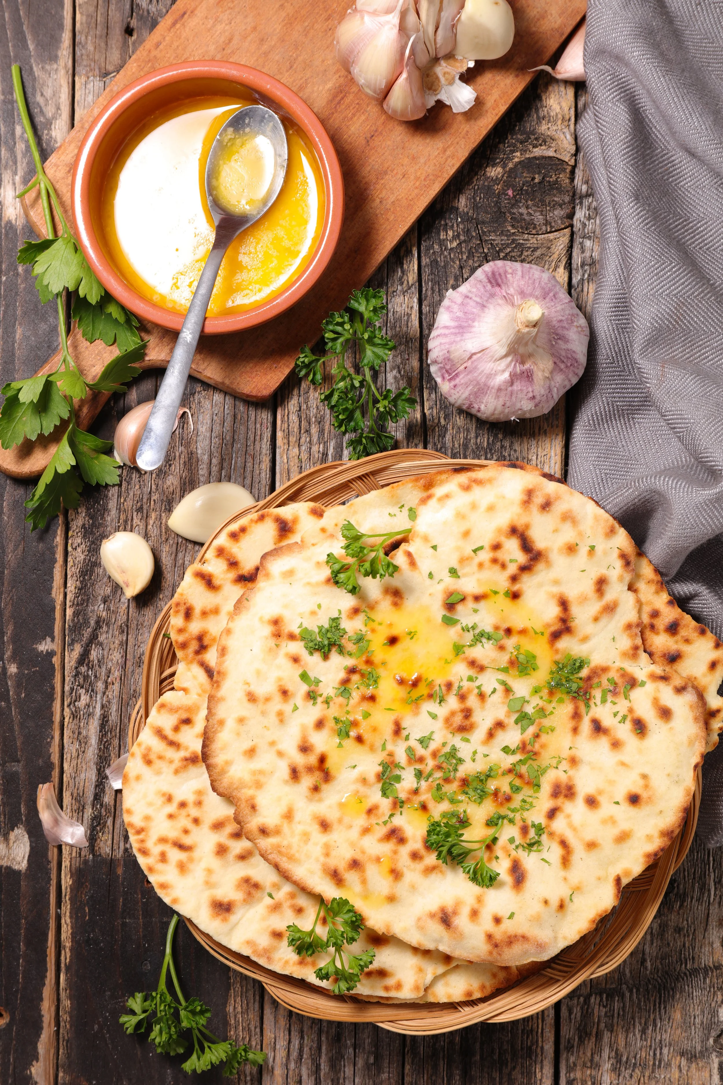 A basket of naan bread garnished with parsley, garlic, and herbs on a rustic wooden table. Behind it, a wooden board holds a bulb of garlic, fresh parsley, and a clay dish with melted butter and garlic.