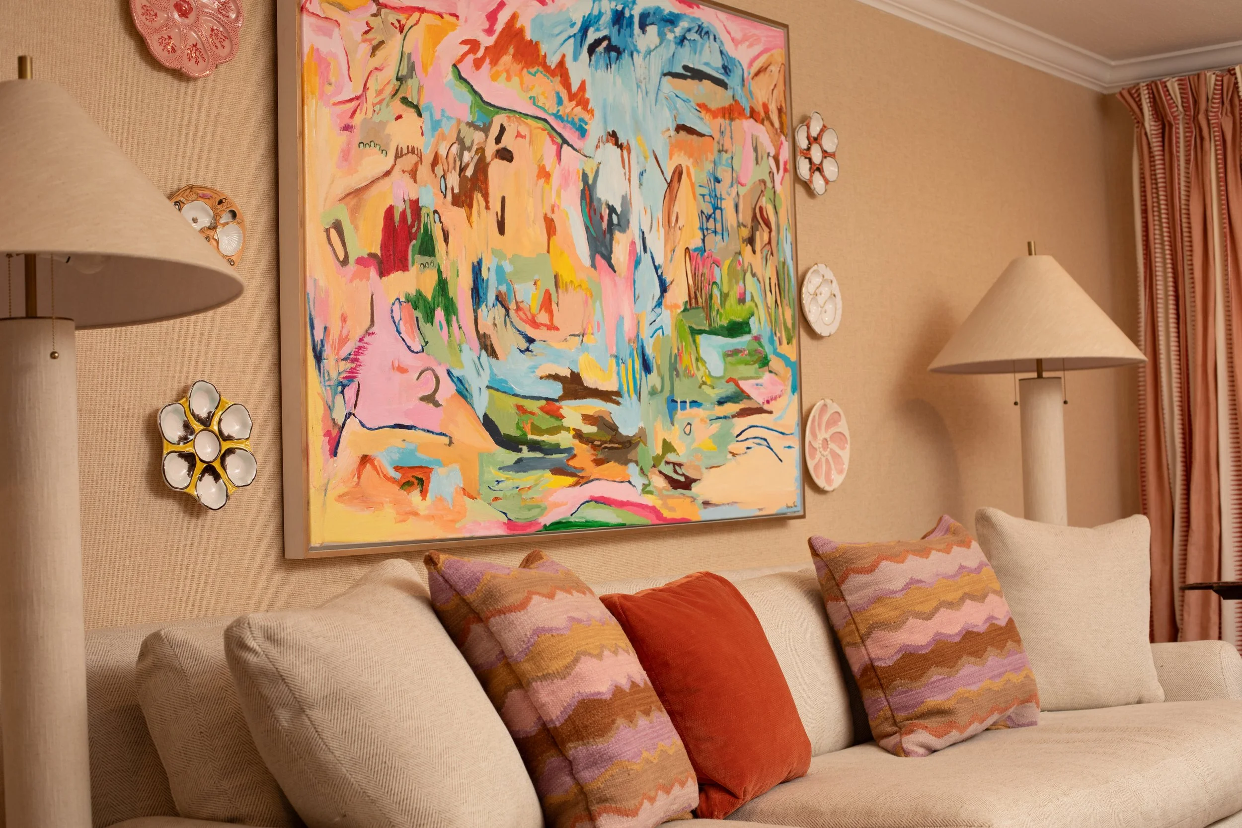 An interior living room wall features a large colorful abstract painting and decorative plates. There are two beige table lamps on either side of the painting, light beige walls, striped curtains, and a beige sofa with multiple patterned throw pillow