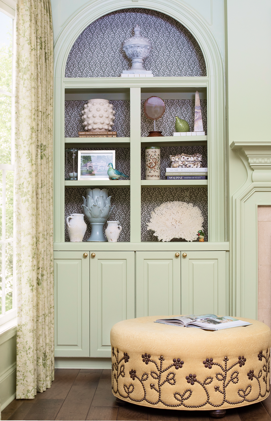 A green built-in bookshelf with decorative items and books, located in a room with light-colored walls and a window with floral curtains. In front of the bookshelf is a round, upholstered ottoman with embroidery and an open magazine on top.