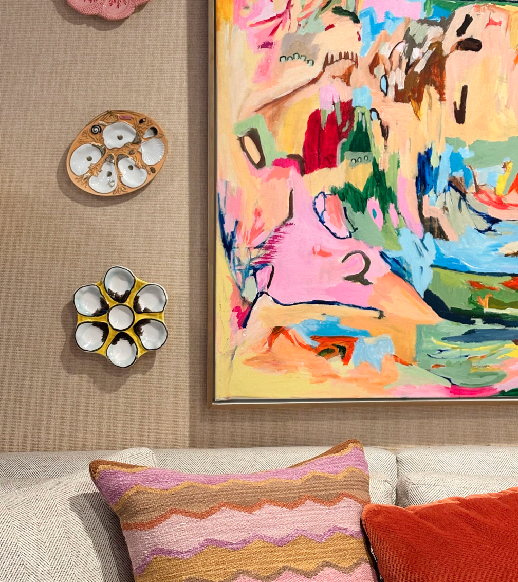 A colorful abstract painting hangs on a beige wall above a white couch with pink, orange, and purple patterned pillows. Two decorative plates, one with seashell shapes and the other with a flower design, are mounted on the wall to the left of the painting.