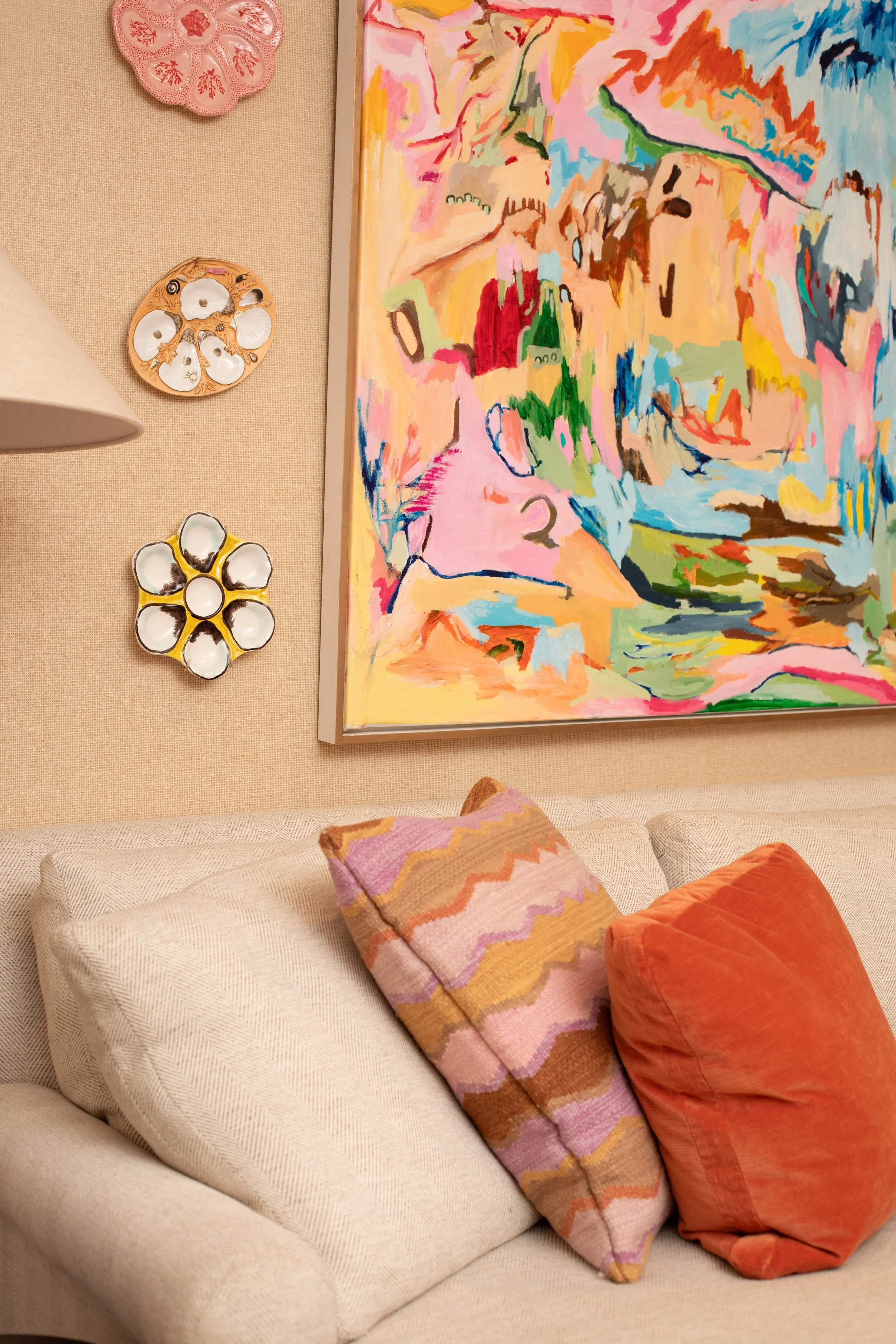 A beige sofa with patterned pink and orange pillows, a colorful abstract painting on the wall, and two decorative sconces, one pink and one green with white shells, above the sofa.