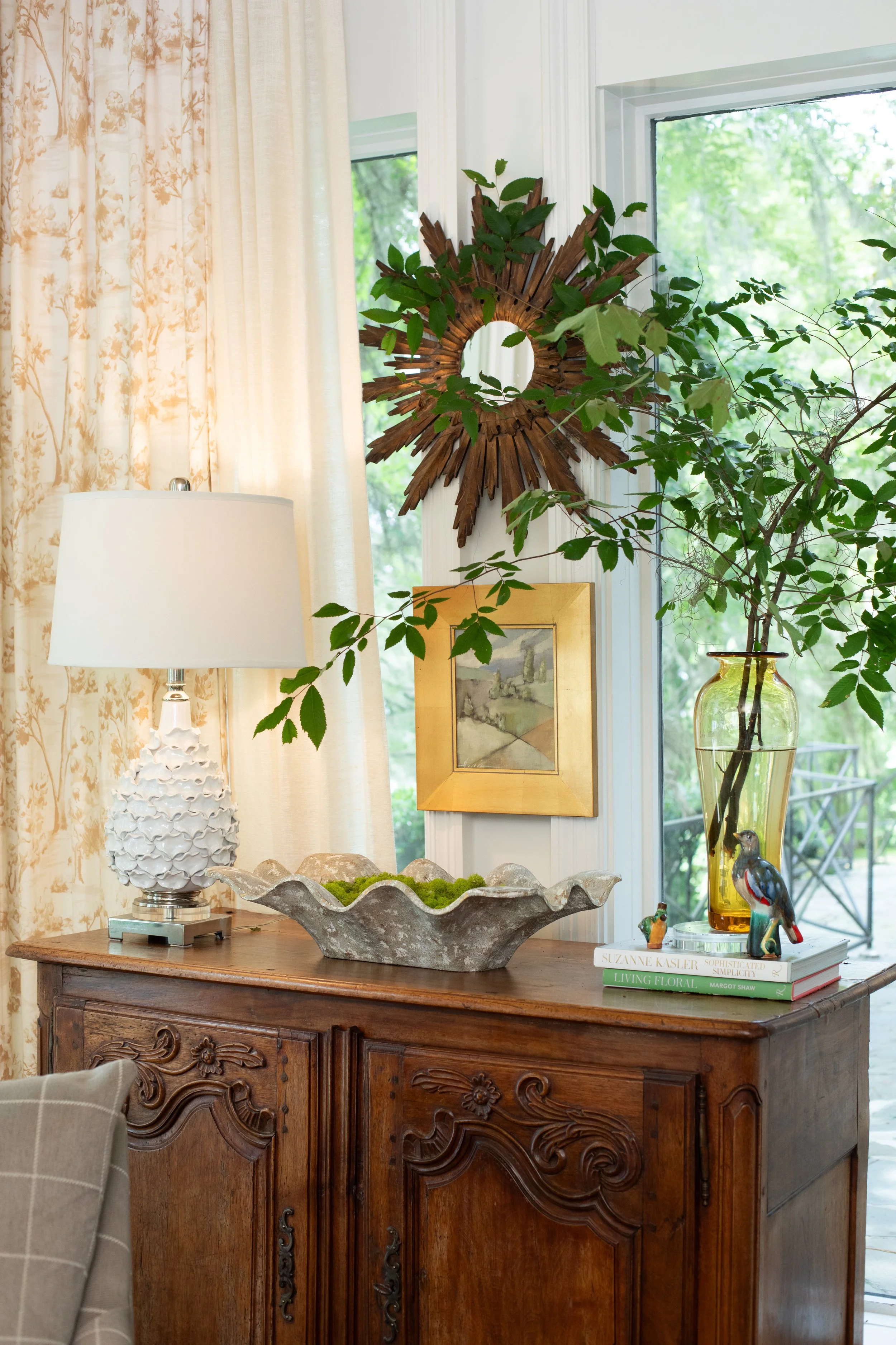 A wooden sideboard with ornate carved details, topped with a decorative vase, a large dish with green moss, a white lamp with a textured base, and a stack of books including "Living Floral," "Simplicity," and others. A window with greenery outside an