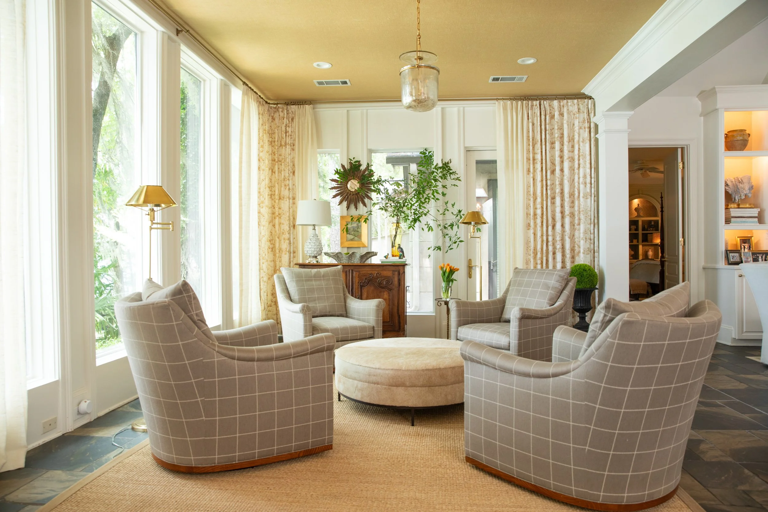 A cozy living room with multiple beige plaid armchairs arranged in a semi-circle around a round ottoman. Large windows with cream curtains allow natural light to fill the room. There are lamps and plants on a wooden sideboard, and a doorway to anothe