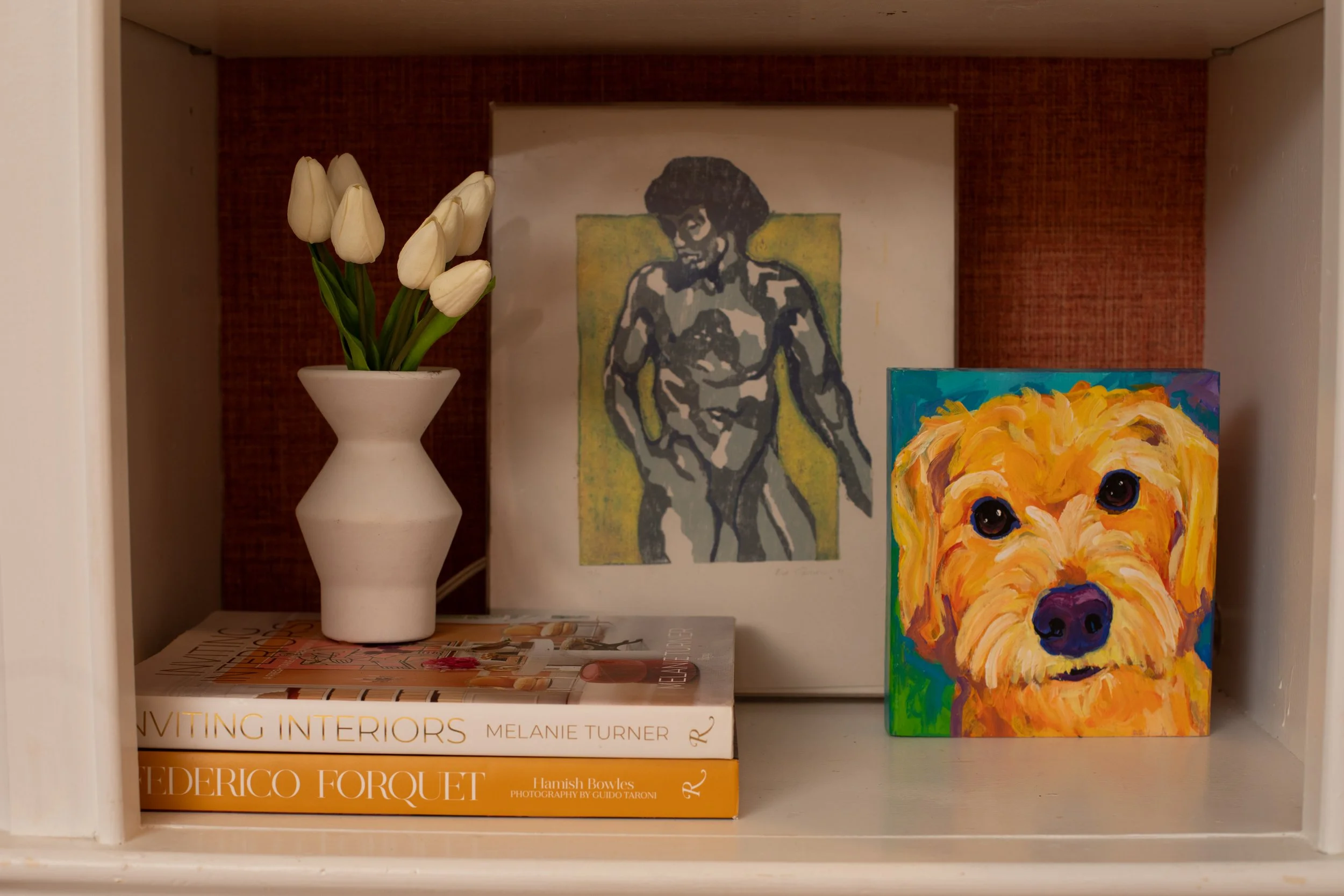 A small white vase with white tulips, a funky tree and woman painting in the background, a colorful dog painting, and a couple of books on interior design and photography on a white shelf.