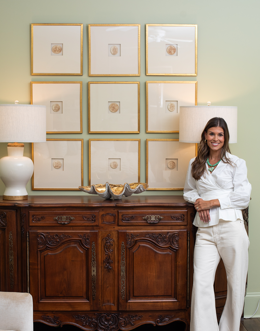 A woman with long brown hair smiling in a white outfit, standing next to a wooden sideboard with lamps and artwork, in a room with green walls.