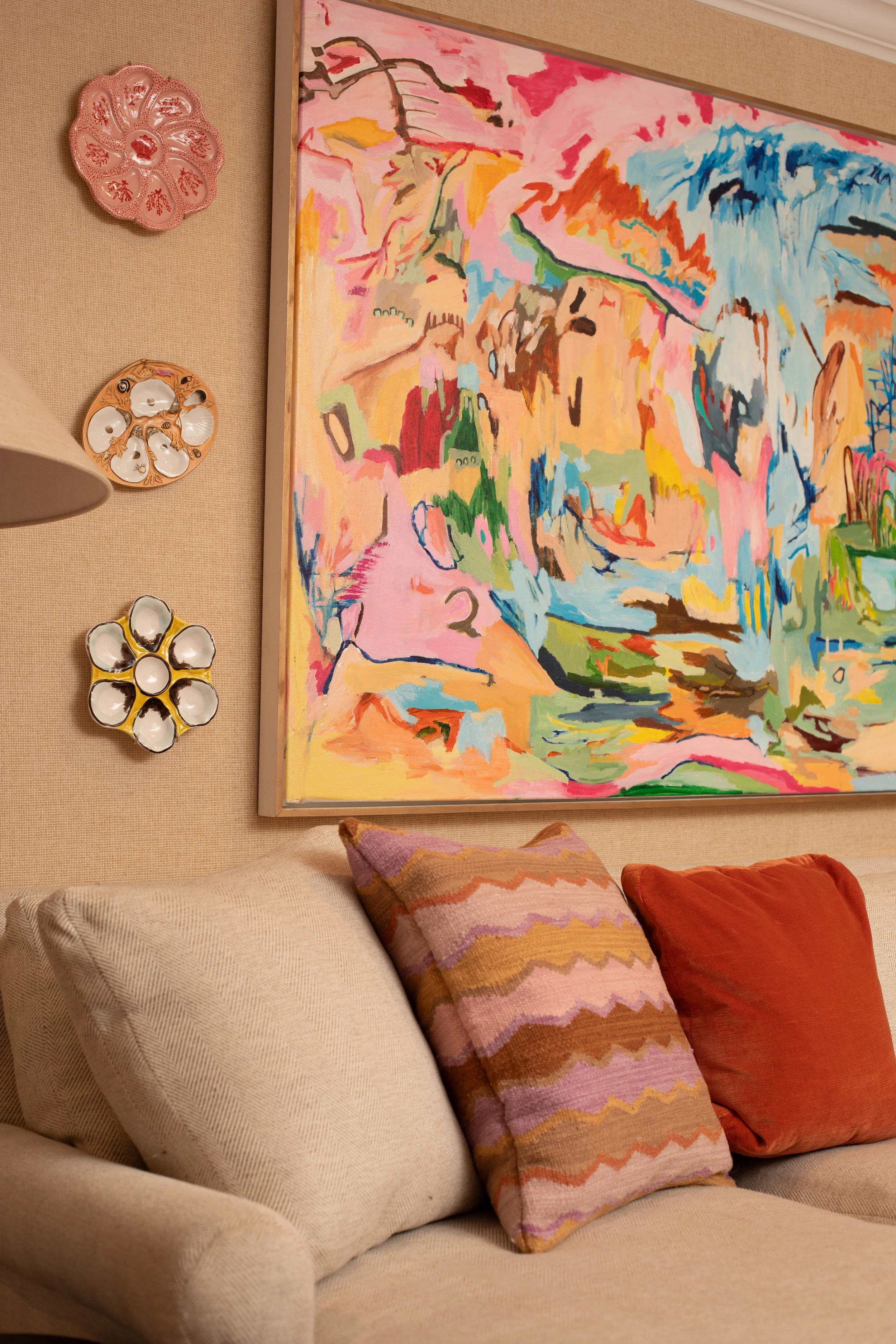 Living room corner featuring a beige sofa with multicolored pillows, a colorful abstract painting on the wall, and decorative wall plates.