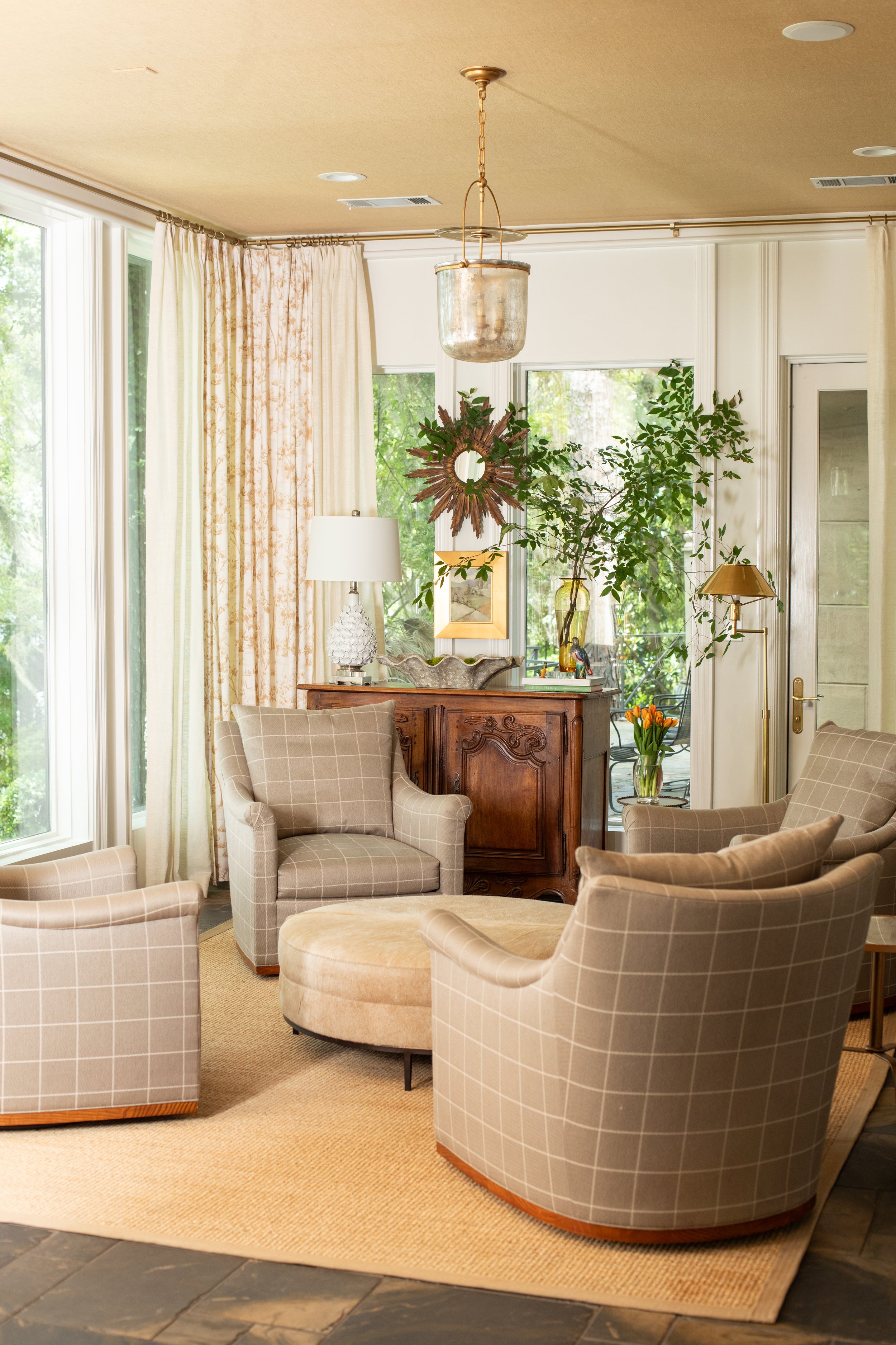 A cozy living room with beige plaid armchairs, a matching ottoman, and a wooden sideboard with a white lamp. Large windows and glass doors let in natural light, with drapes and greenery outside. Decor includes a sunburst mirror, a large vase with gre