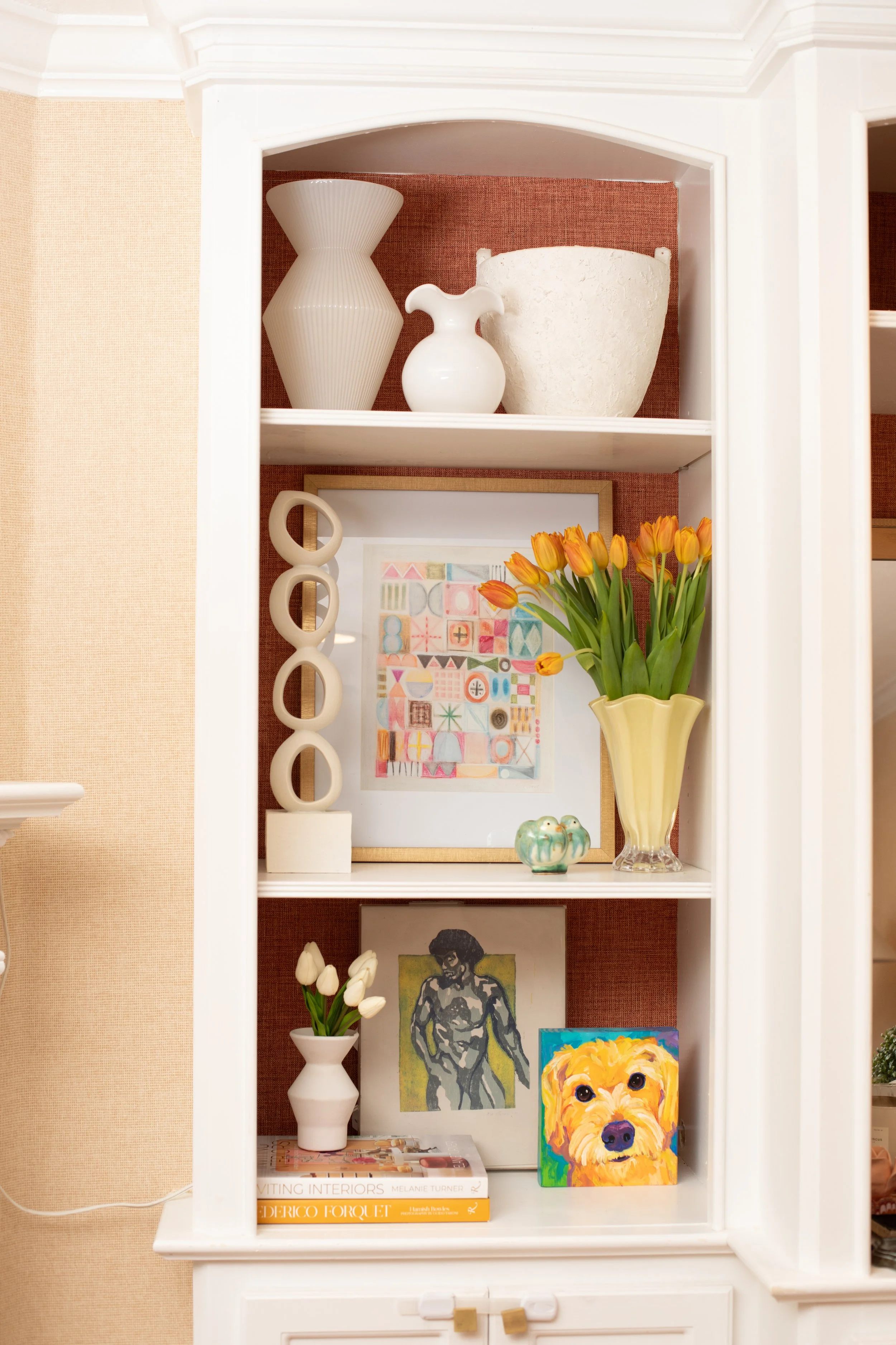Decorative white built-in bookshelf with three shelves, holding vases, framed artwork, books, a small sculpture, and colorful tulips in a yellow vase, against a textured brown wall.