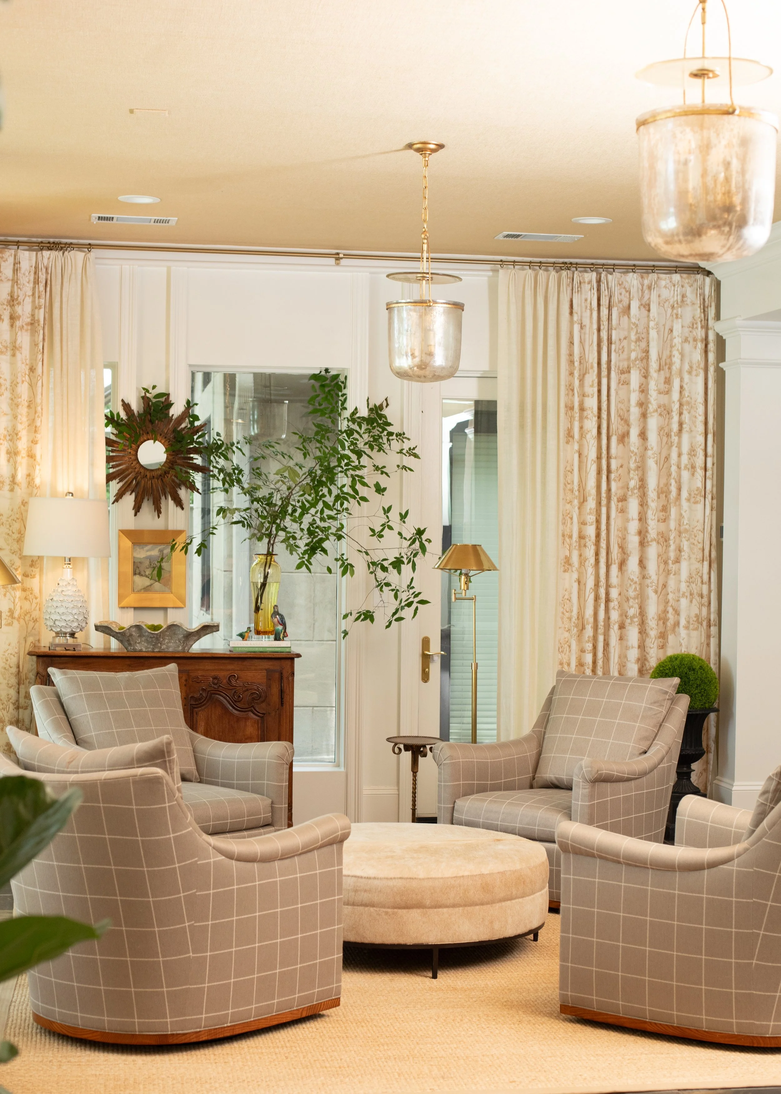 A cozy living room with five plaid armchairs surrounding a round beige ottoman, a wooden sideboard with decor, a leafy green plant, and windows with cream-colored curtains.