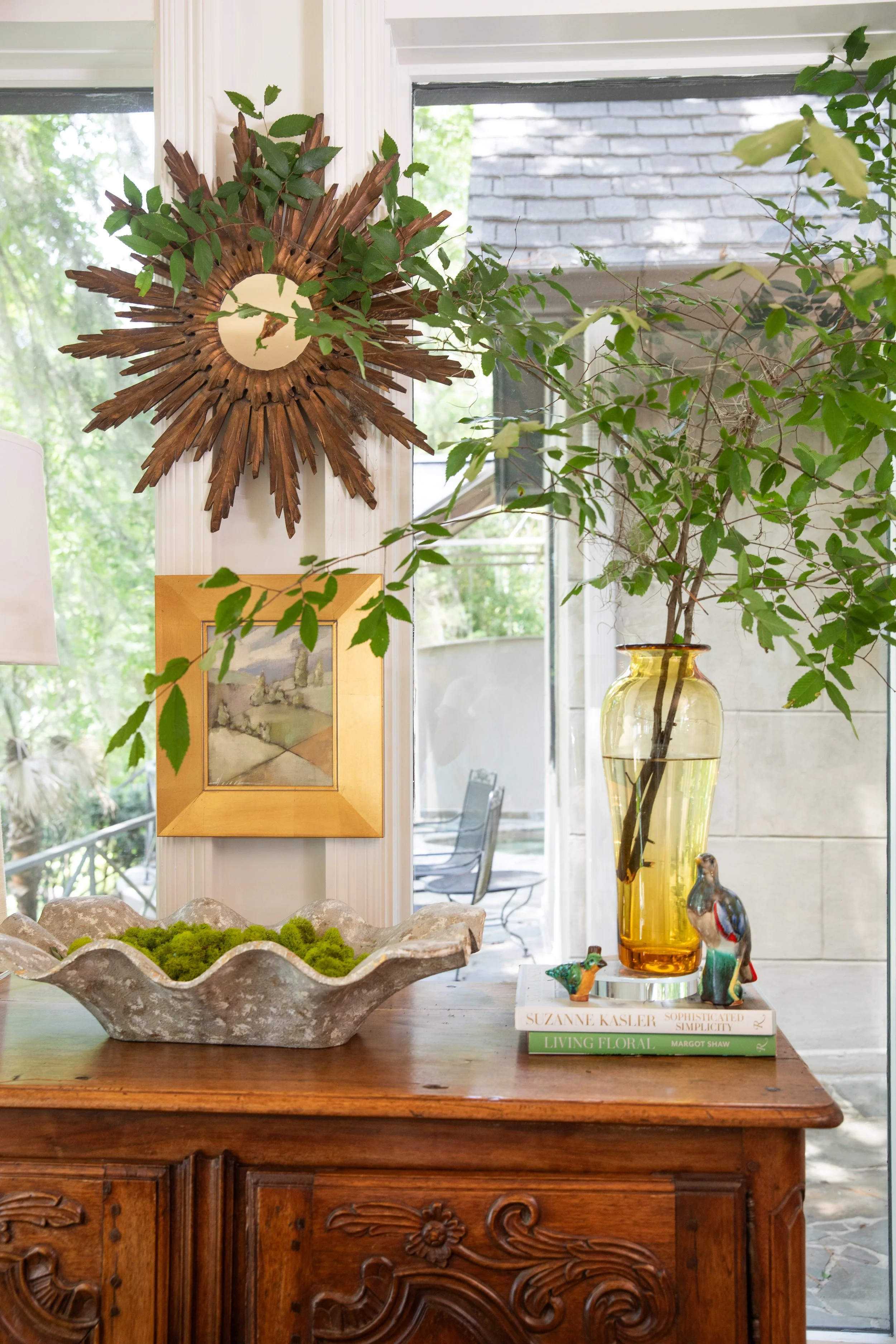 A wooden sideboard with ornate carvings holding decorative objects including a ceramic dish with moss, books, a yellow glass vase with branches, and ceramic bird figurines. In the background, there is a window and framed artwork.