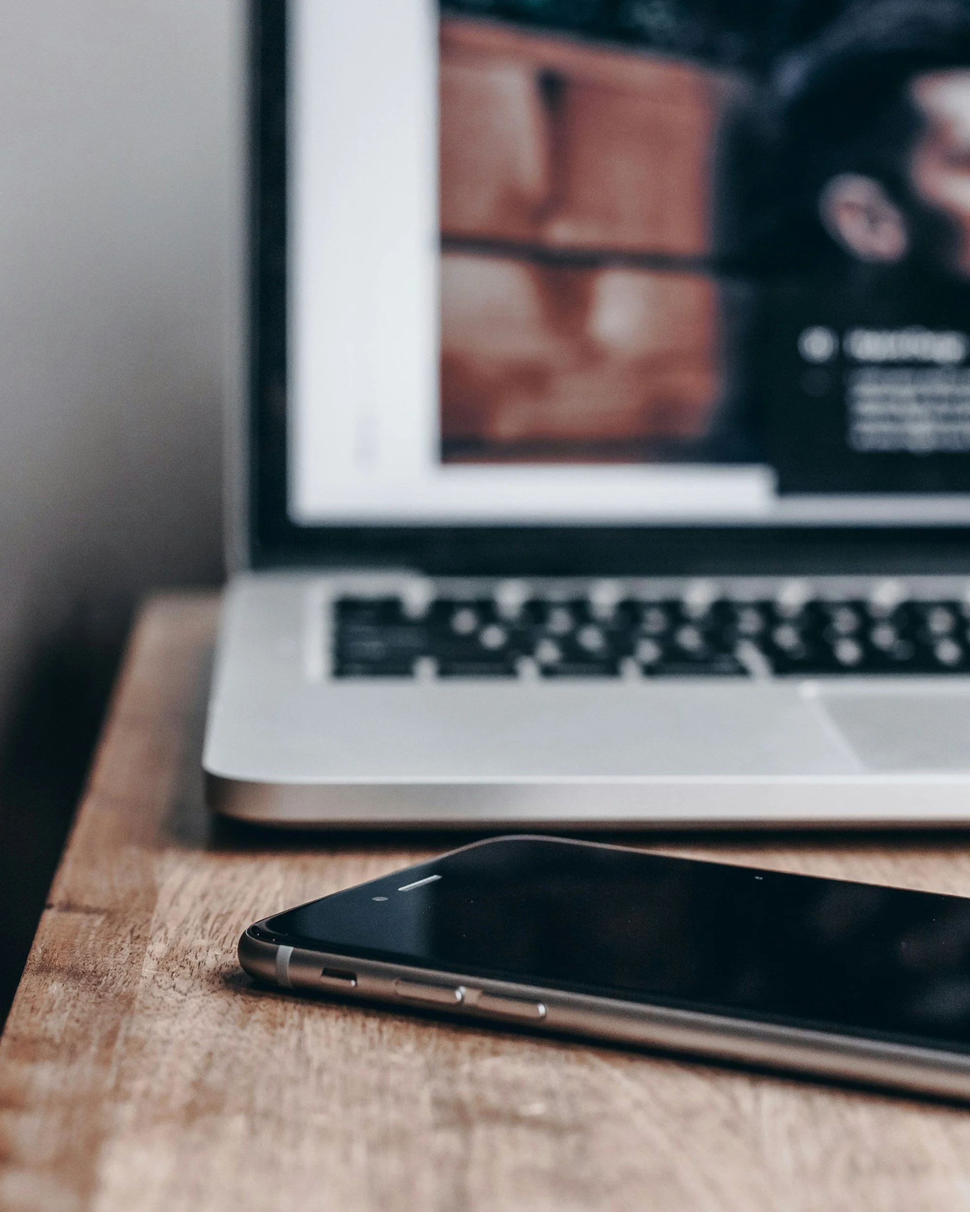 Close-up of a smartphone lying on a wooden surface with a laptop and a blurred bookshelf in the background.