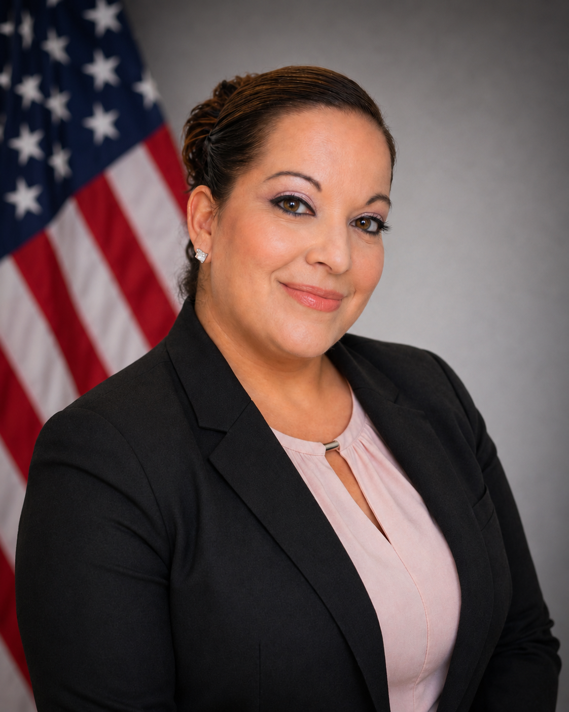 A woman in a black blazer and pink blouse, smiling, with the American flag in the background.
