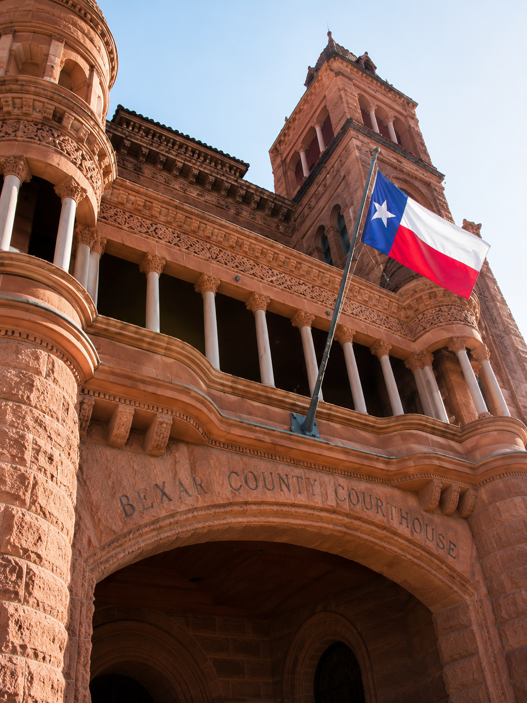 View of Bexar County Courthouse in San Antonio, Texas, showing a red stone building with arches and a tower, with a Texas flag flying outside.