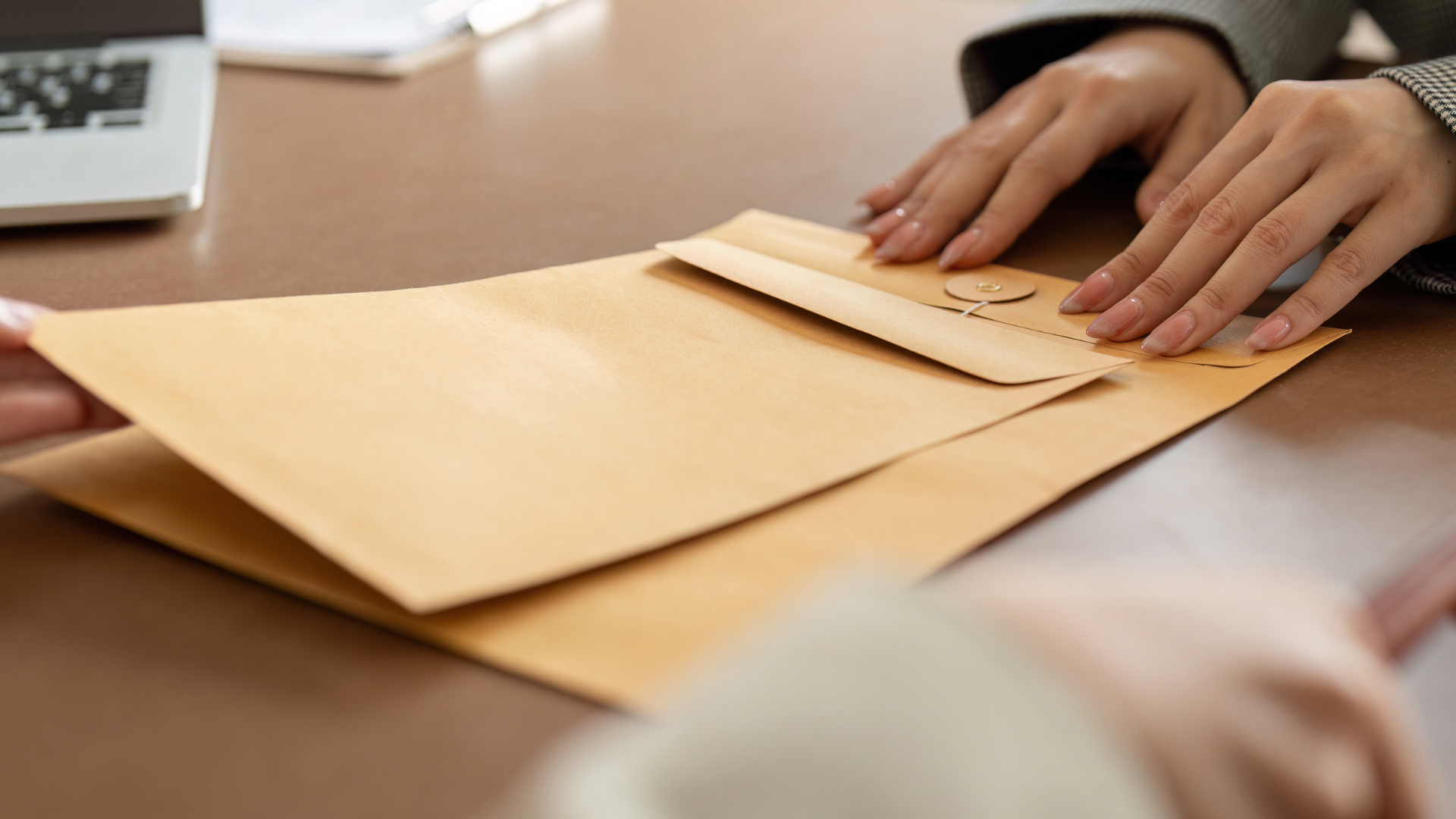 Person using a measuring tape to measure a large envelope on a desk, with a laptop in the background.