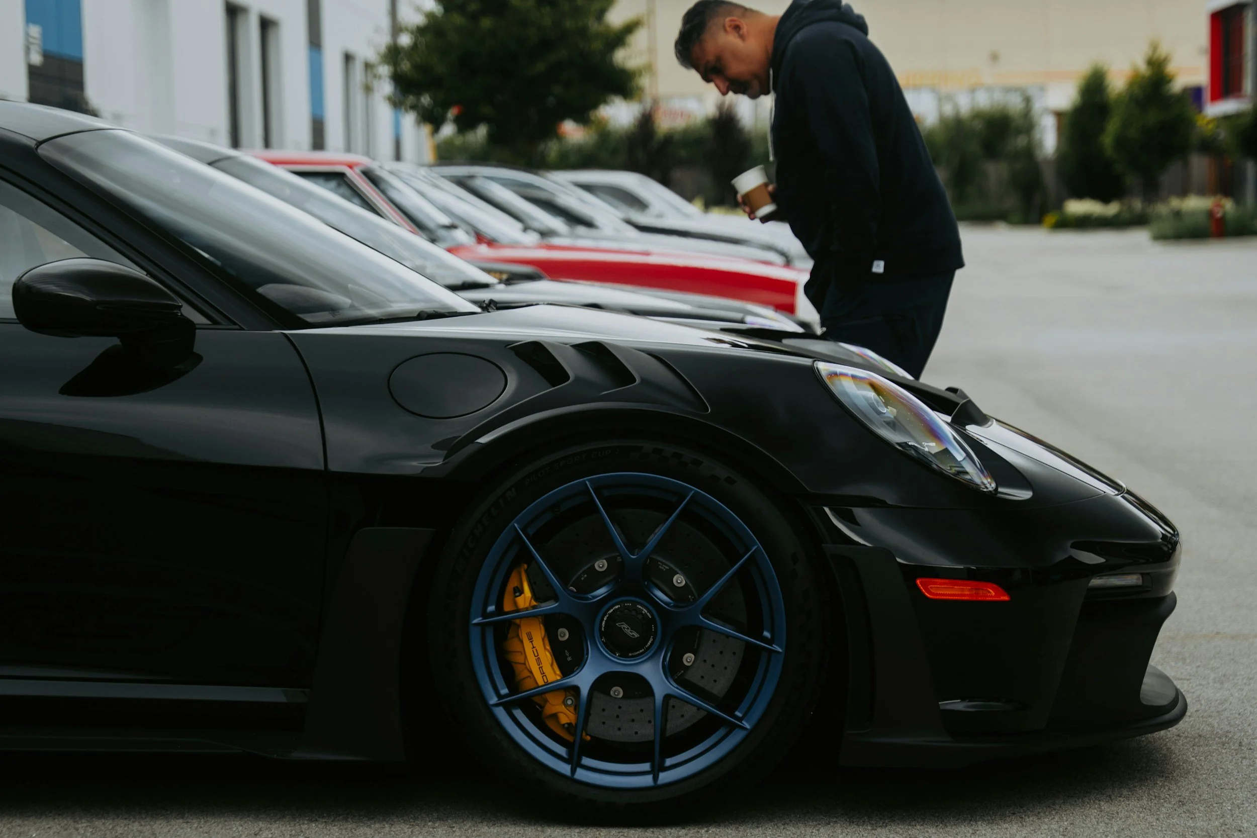 A man in a black hoodie examining a black sports car and holding a coffee cup in an outdoor parking lot lined with other cars and trees.