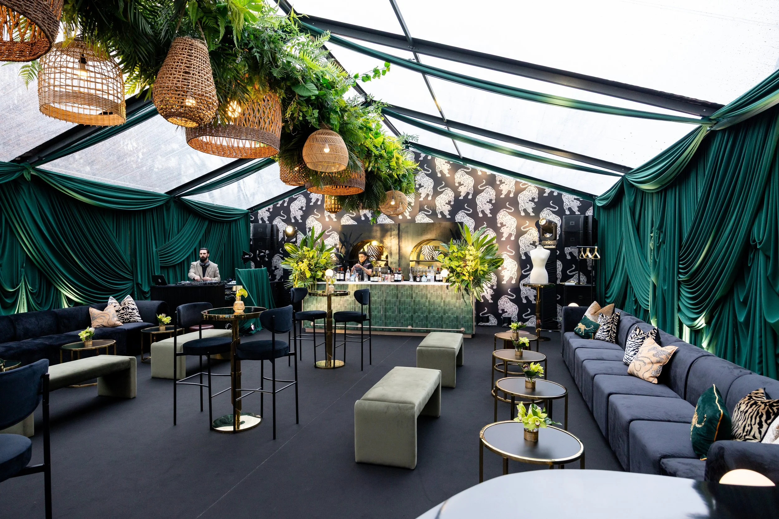 Interior of a decorated event venue with green draped fabric walls, hanging wicker lamps, lush green foliage ceiling decor, a bar with floral arrangements, modern seating with velvet couches and small tables, and a patterned wall with white elephants.