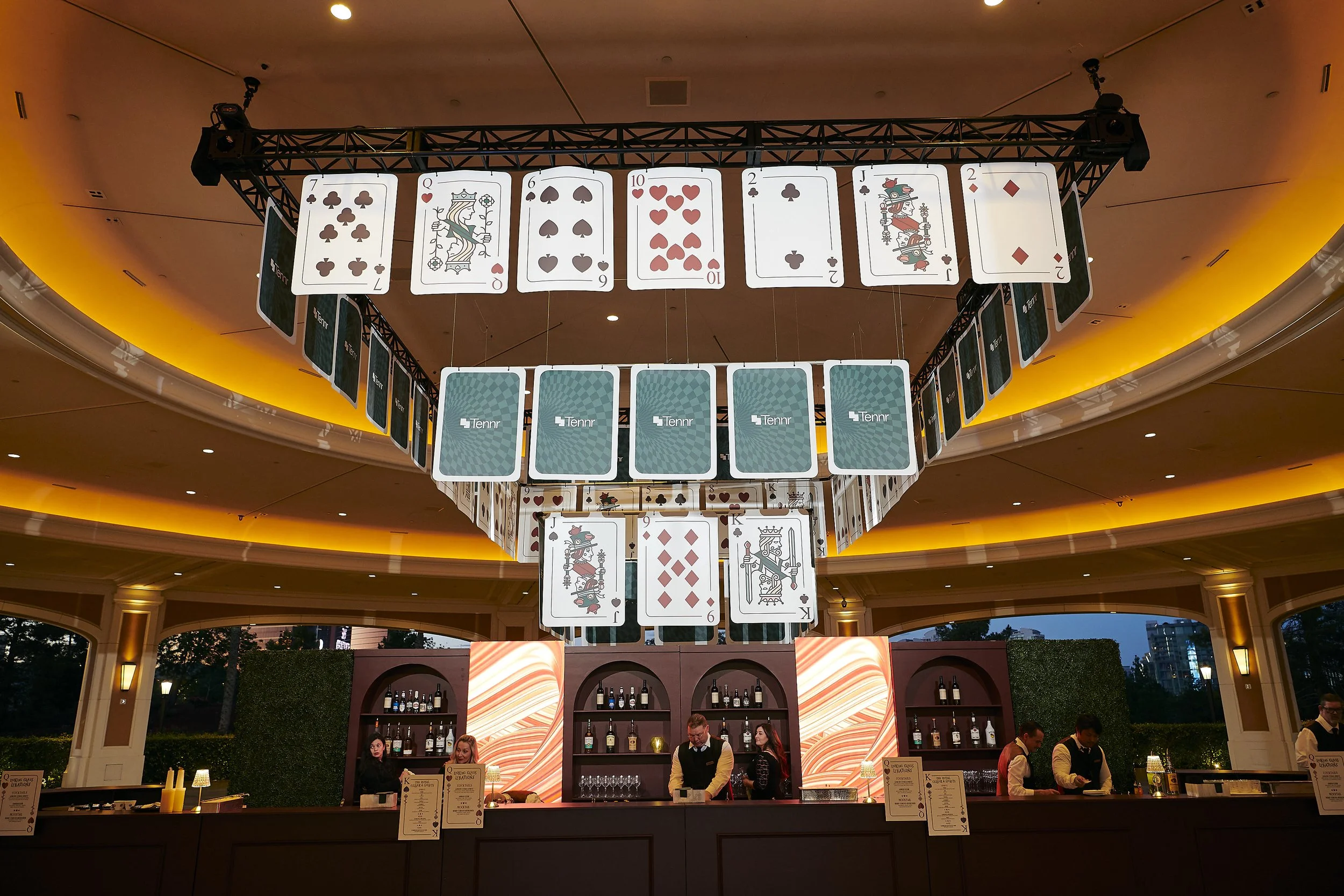 Large hanging display of playing cards above a bar with bartenders and patrons in a casino or event setting, illuminated with warm lighting.