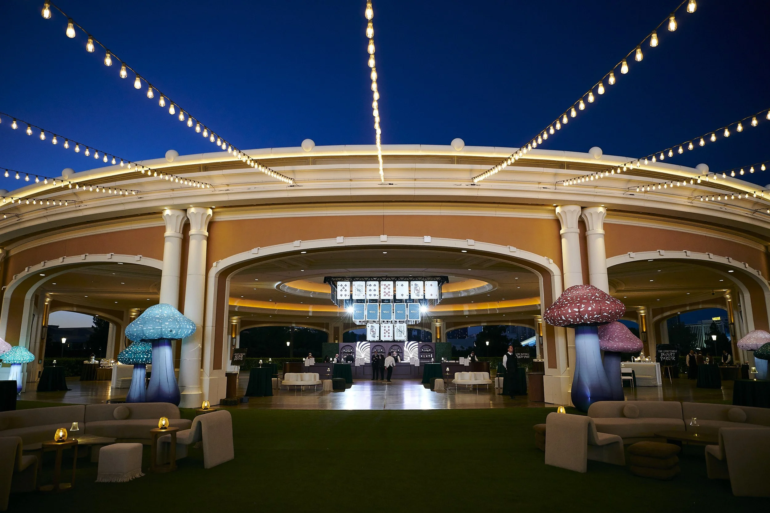 Night view of an elegant outdoor event space with string lights, whimsical mushroom-shaped decorations, and a bar area underneath a large arching structure.
