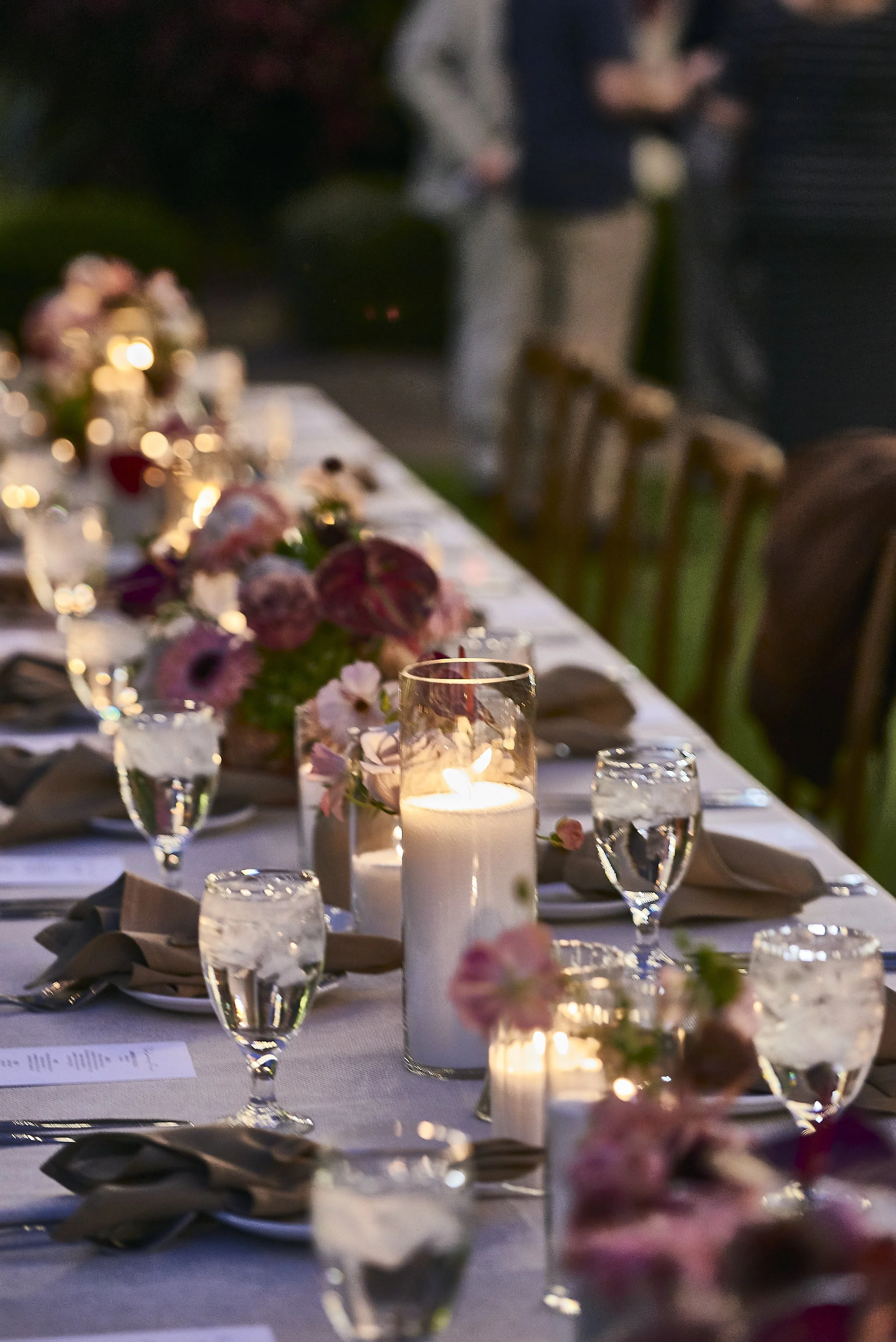 A dinner table set for an event at dusk with candles, flowers, wine glasses, silverware, and plates. The background shows blurred people standing and socializing outdoors.