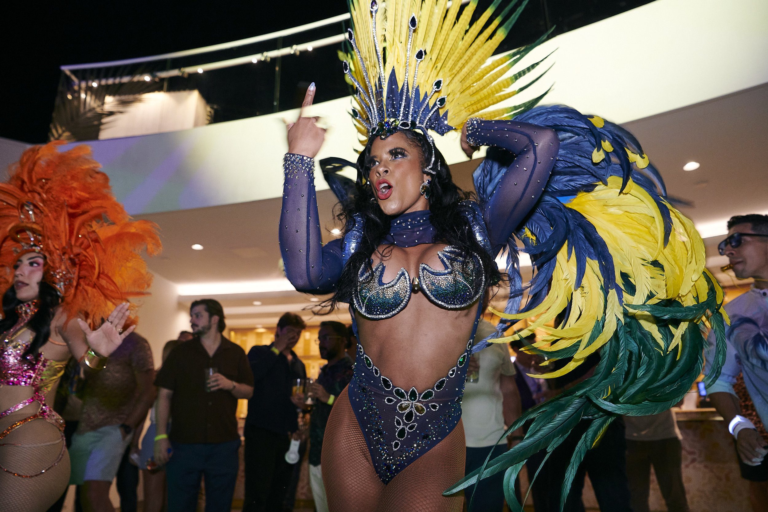 A woman dressed in an ornate, colorful carnival costume with a large yellow and green feathered headdress and matching feathered accessories. She is dancing or performing at an indoor event, with other performers and spectators in the background.