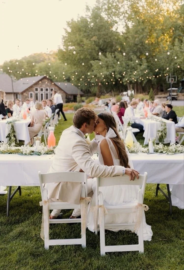 A newlywed couple kissing at their outdoor wedding reception, surrounded by guests seated at decorated tables with string lights overhead, in a lush garden setting with trees and a house in the background.