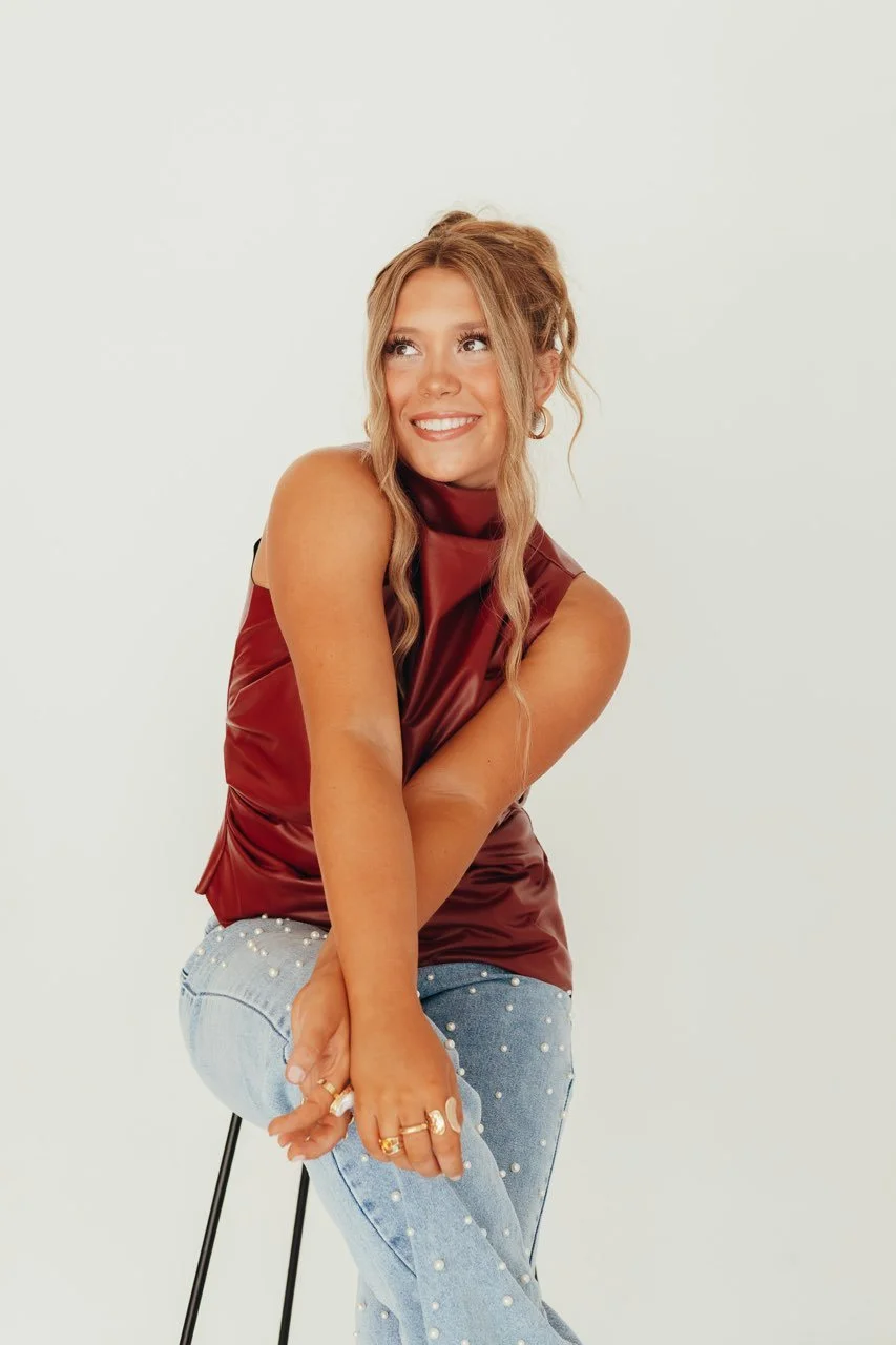 A young woman with long wavy hair sitting on a stool, smiling and looking to the side, wearing a sleeveless maroon satin top and light blue jeans decorated with pearls.