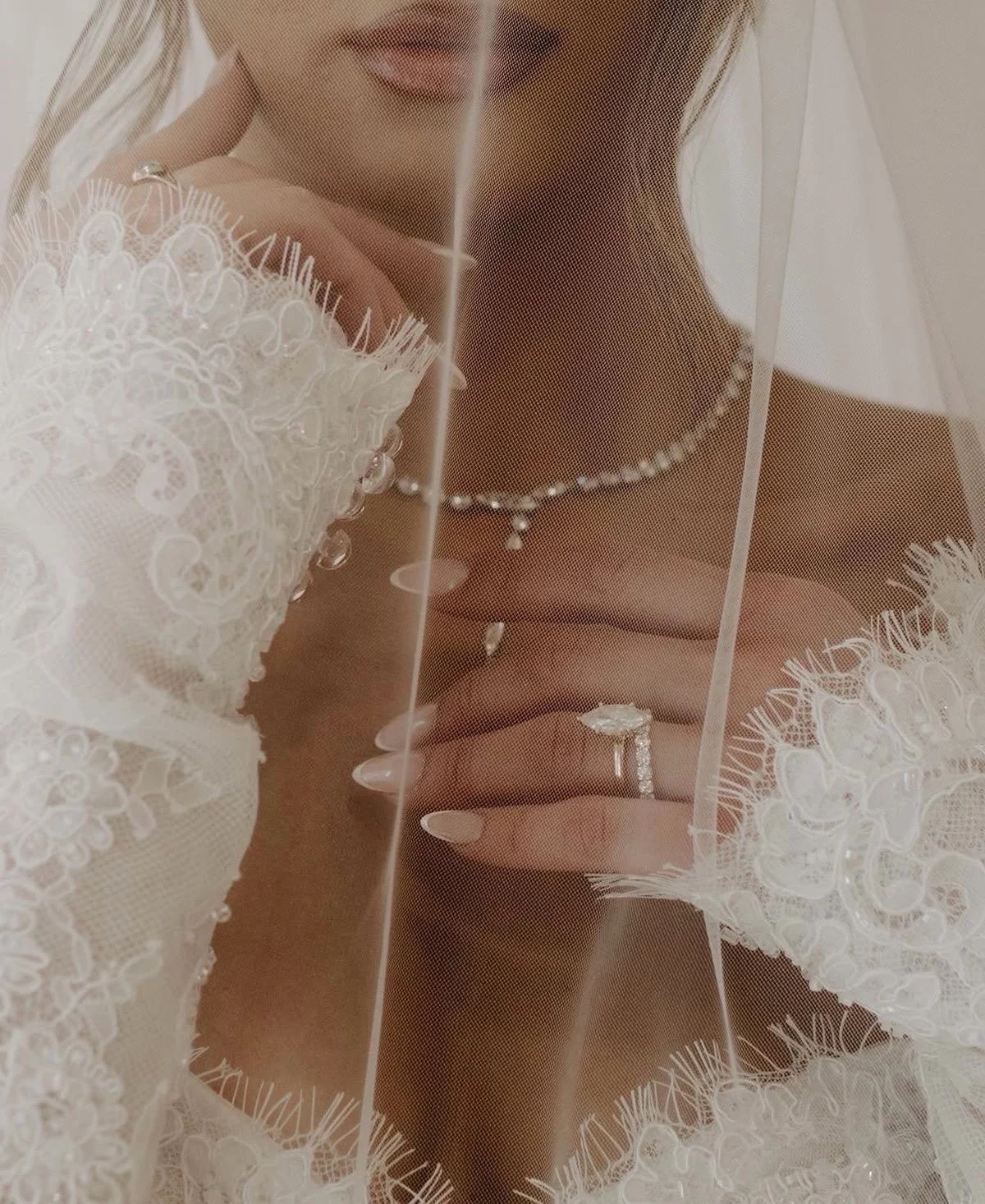 Close-up of a bride showing her wedding ring, necklace, and dress, seen through a sheer veil or fabric with embroidered lace.